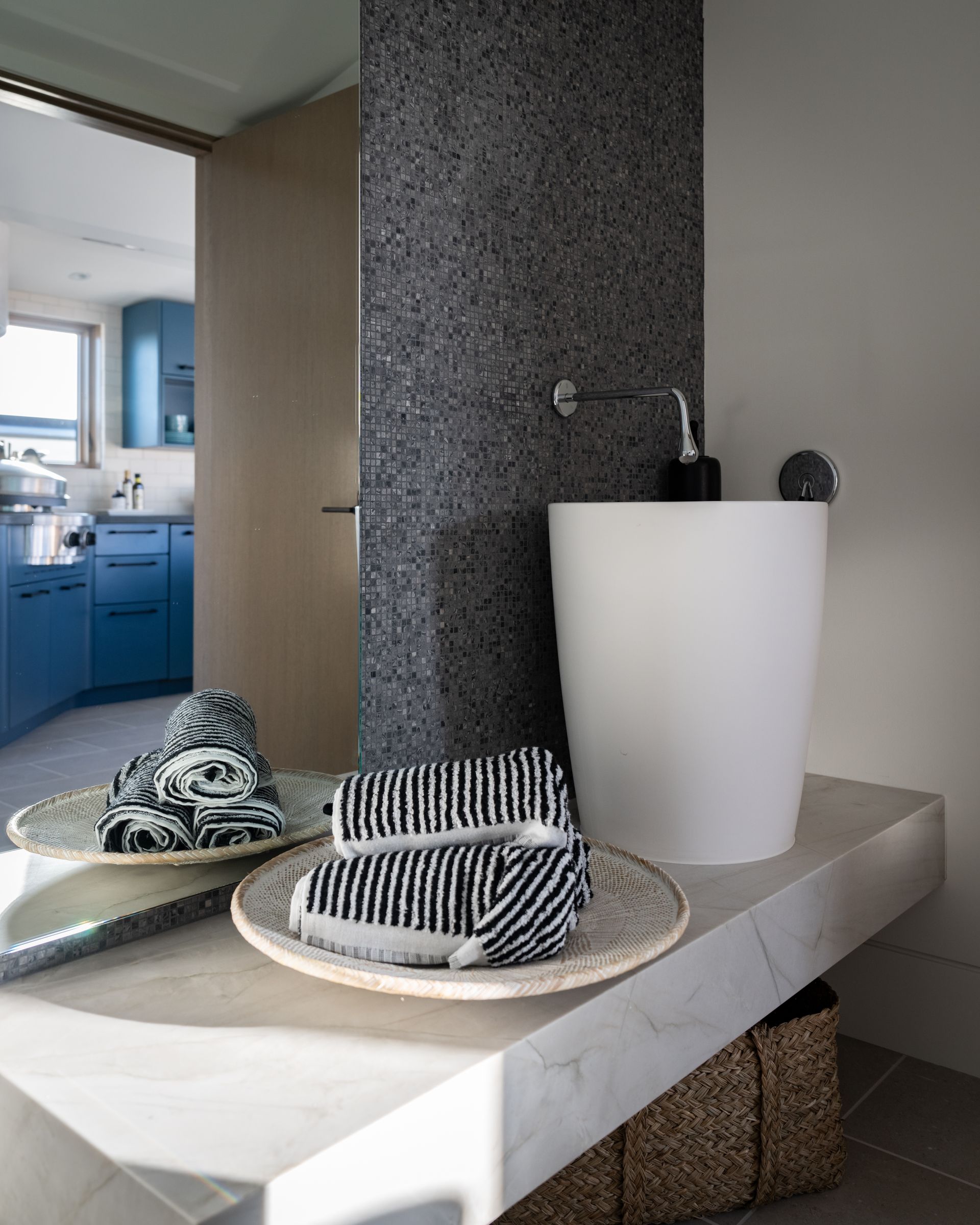 A white pedestal sink sits on a stone vanity next to a dark textured wall, with patterned towels resting on wicker trays.