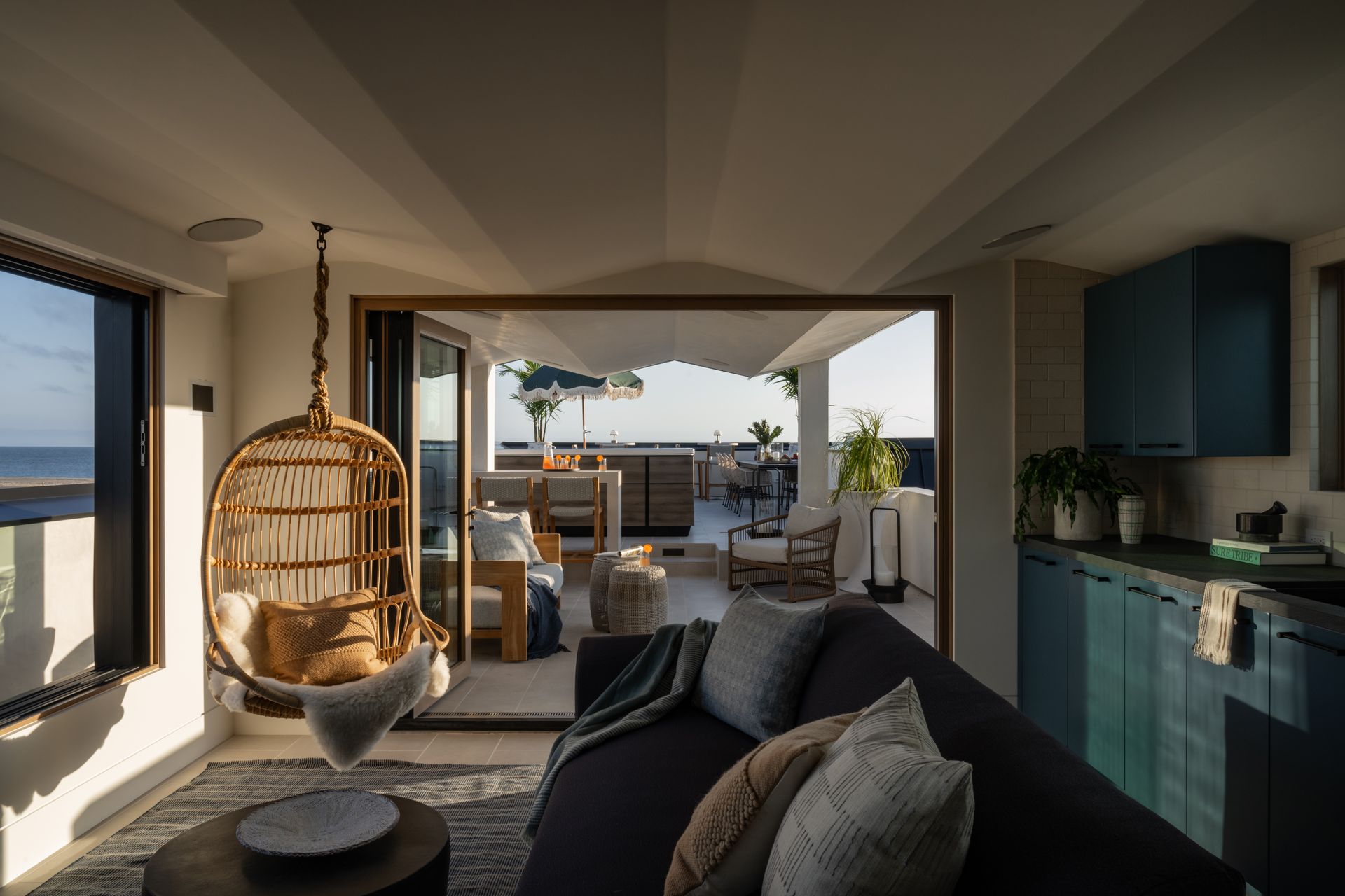 Modern coastal living space with a hanging wicker chair, dark sofa, and open balcony views.