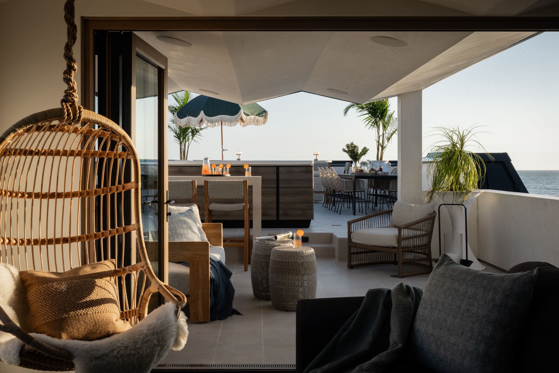 A sunlit terrace with a wicker swing chair, outdoor dining area, lounge seating, and a view of the ocean.