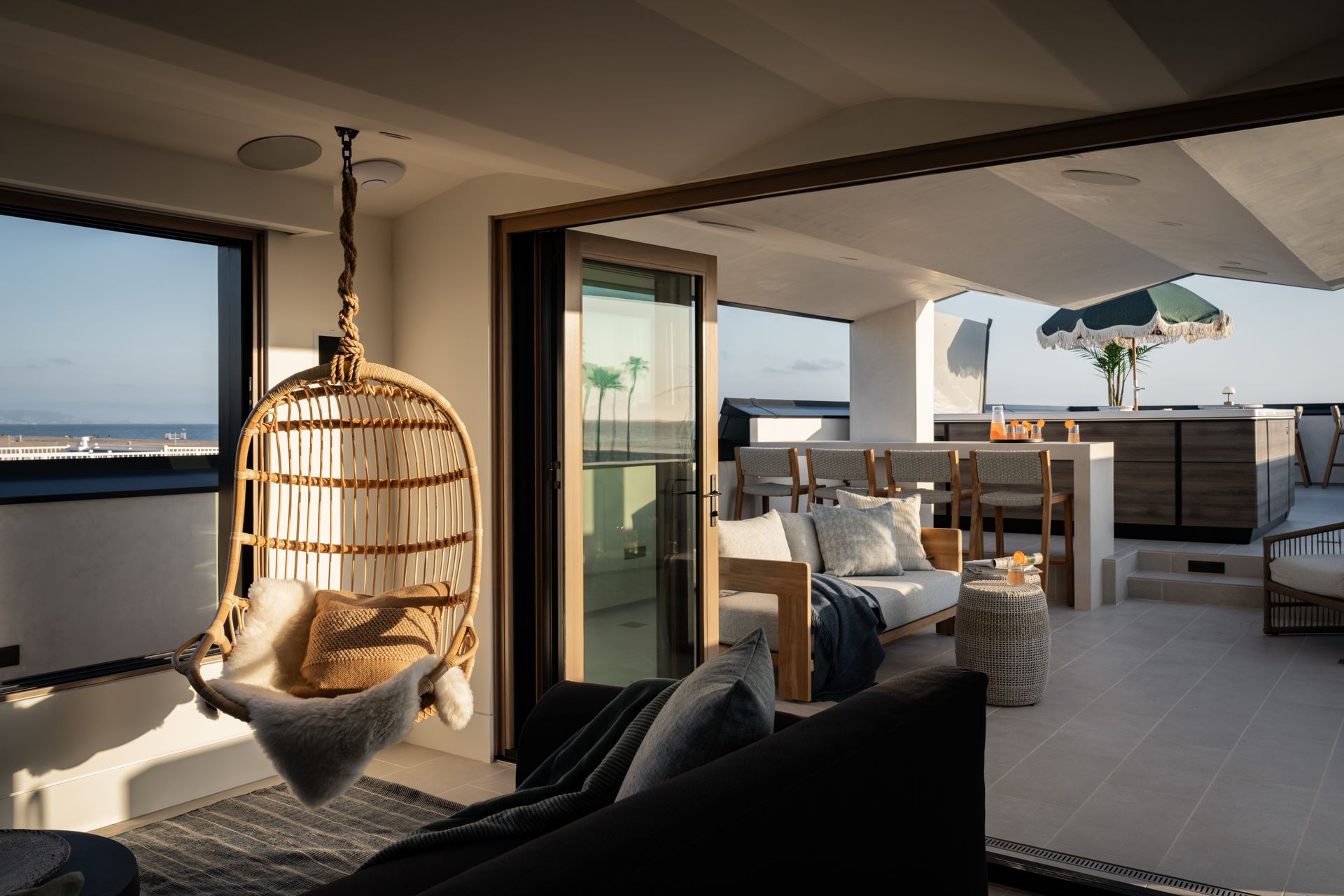 A modern living room with a hanging rattan chair, opening onto a sunlit terrace featuring a bar, lounge area, and ocean view.
