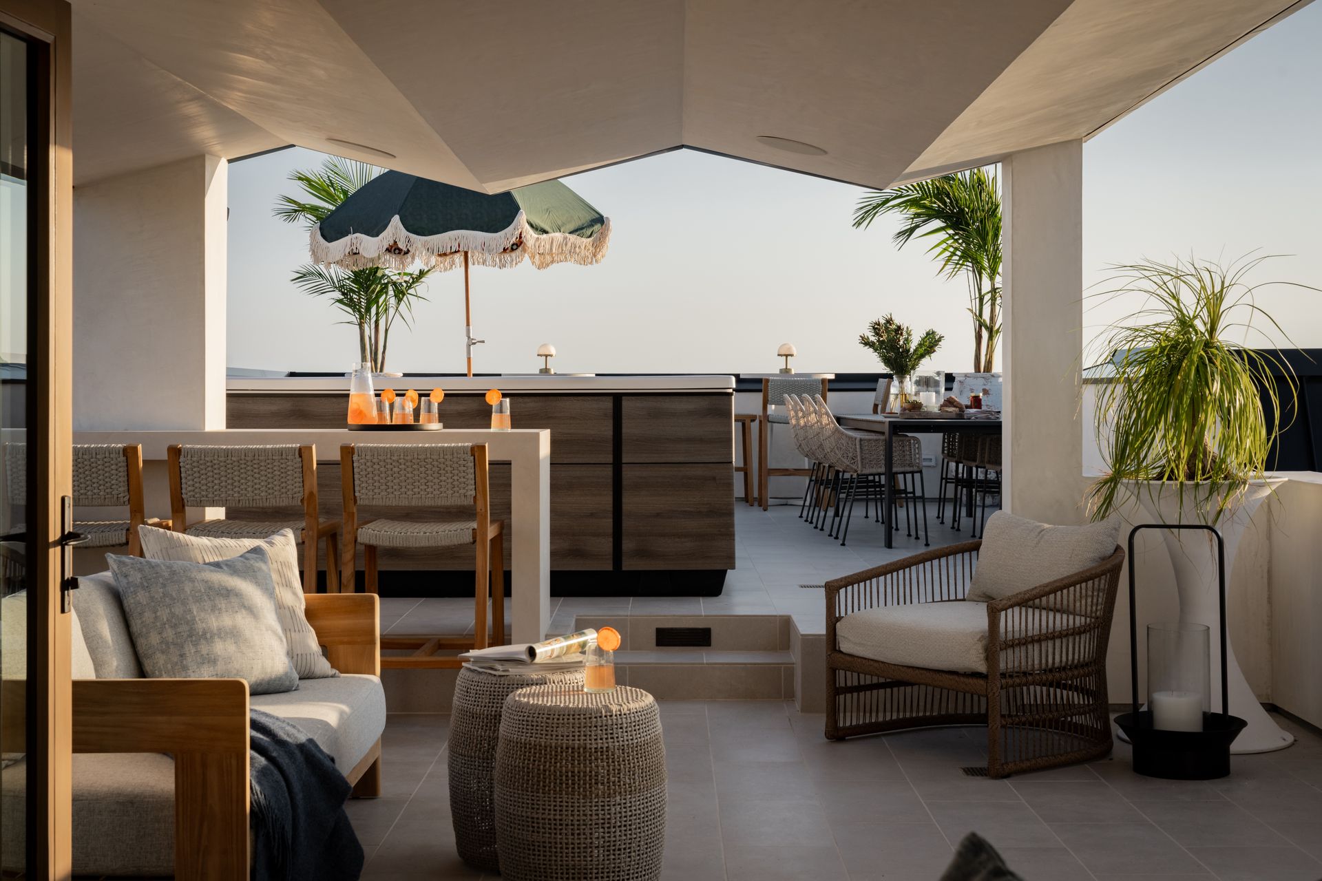 A chic outdoor terrace with neutral-toned seating, a bar area under an umbrella, and potted plants at sunset.