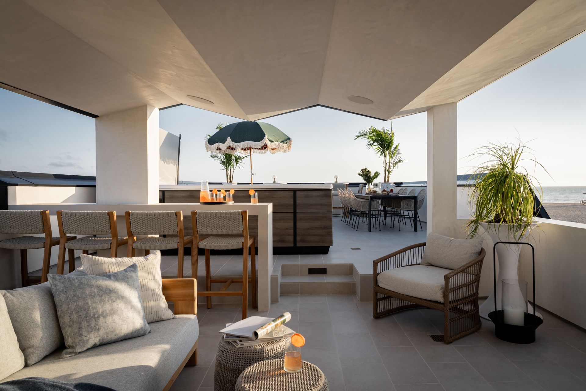 A modern, neutral-toned outdoor terrace with a lounge area, bar seating, and a dining space overlooking the ocean.