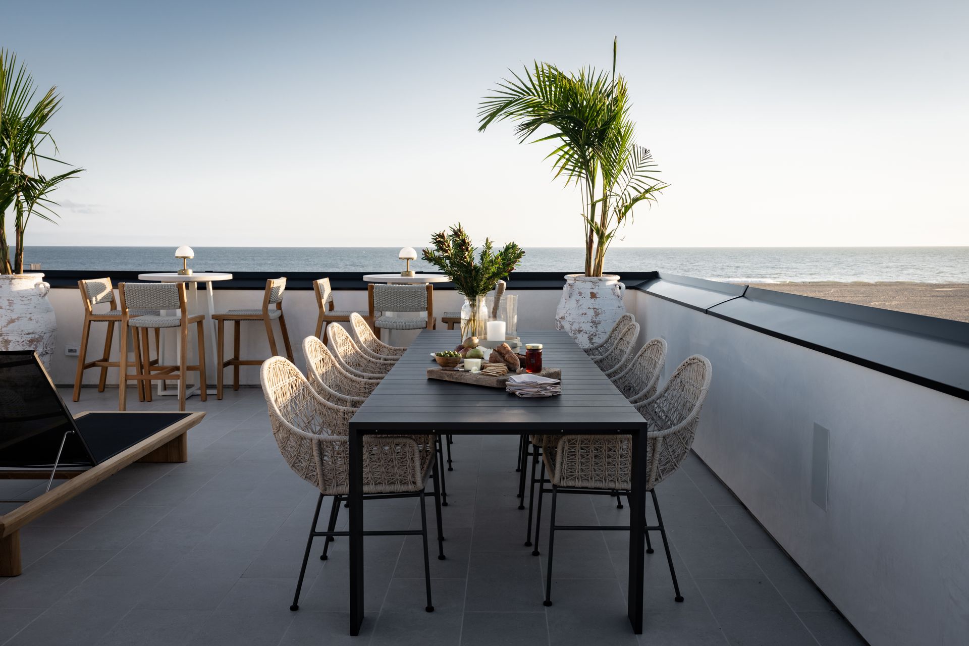 A sleek, long dining table with woven chairs on an outdoor balcony overlooking the ocean at sunset.