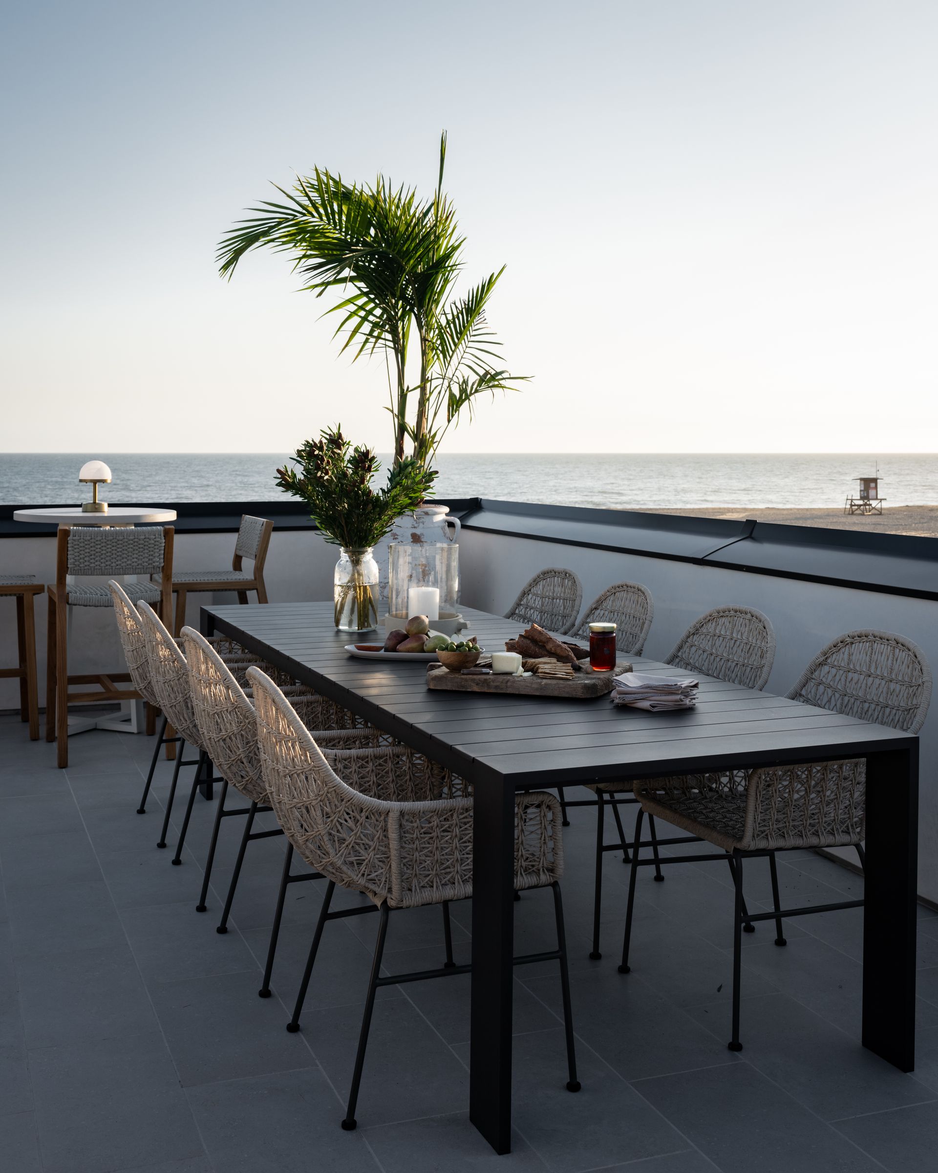 A long outdoor dining table set with food and a tall plant overlooks the ocean from a rooftop deck.