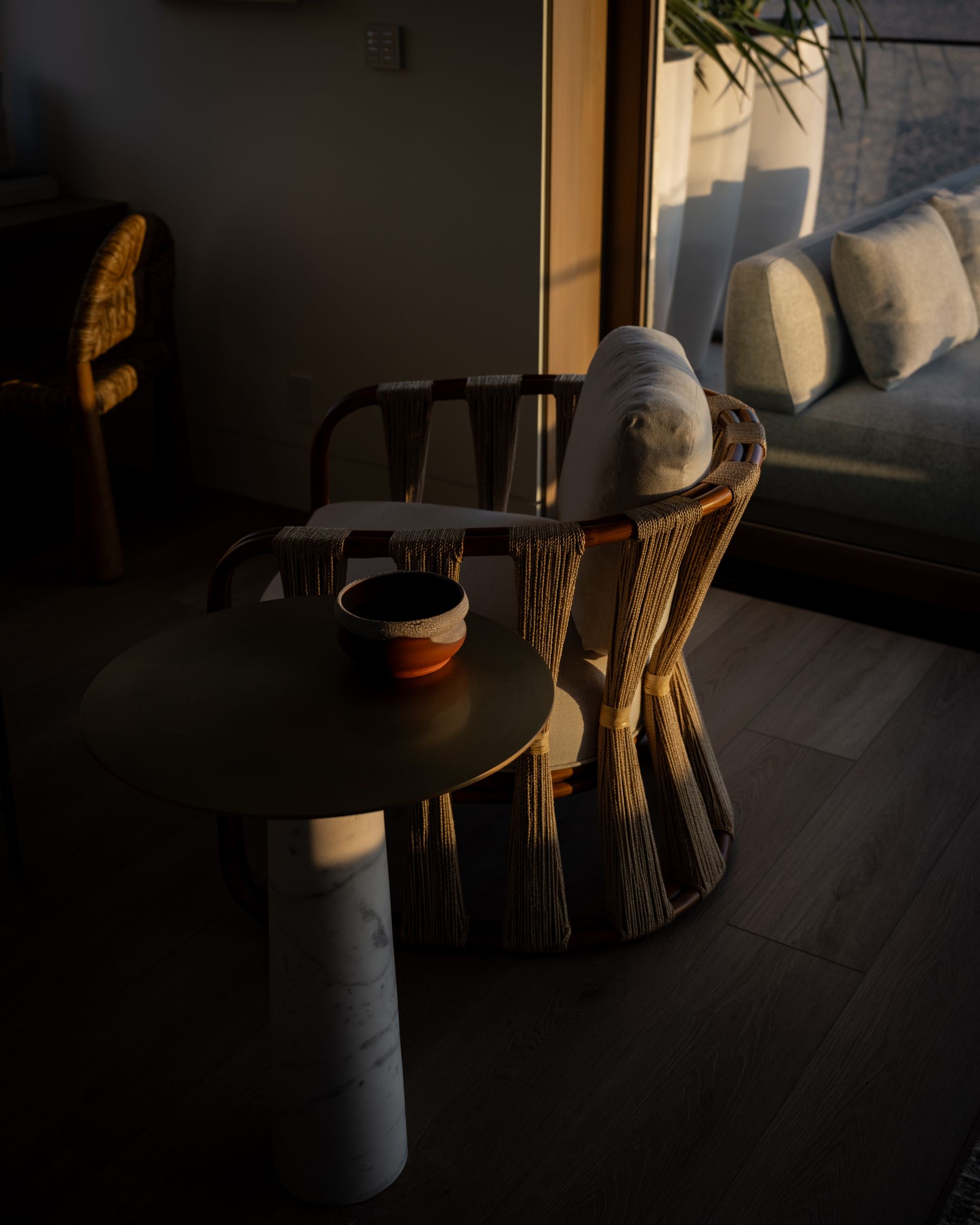 A woven lounge chair and a round side table with a small bowl, illuminated by warm sunlight in a modern living space.