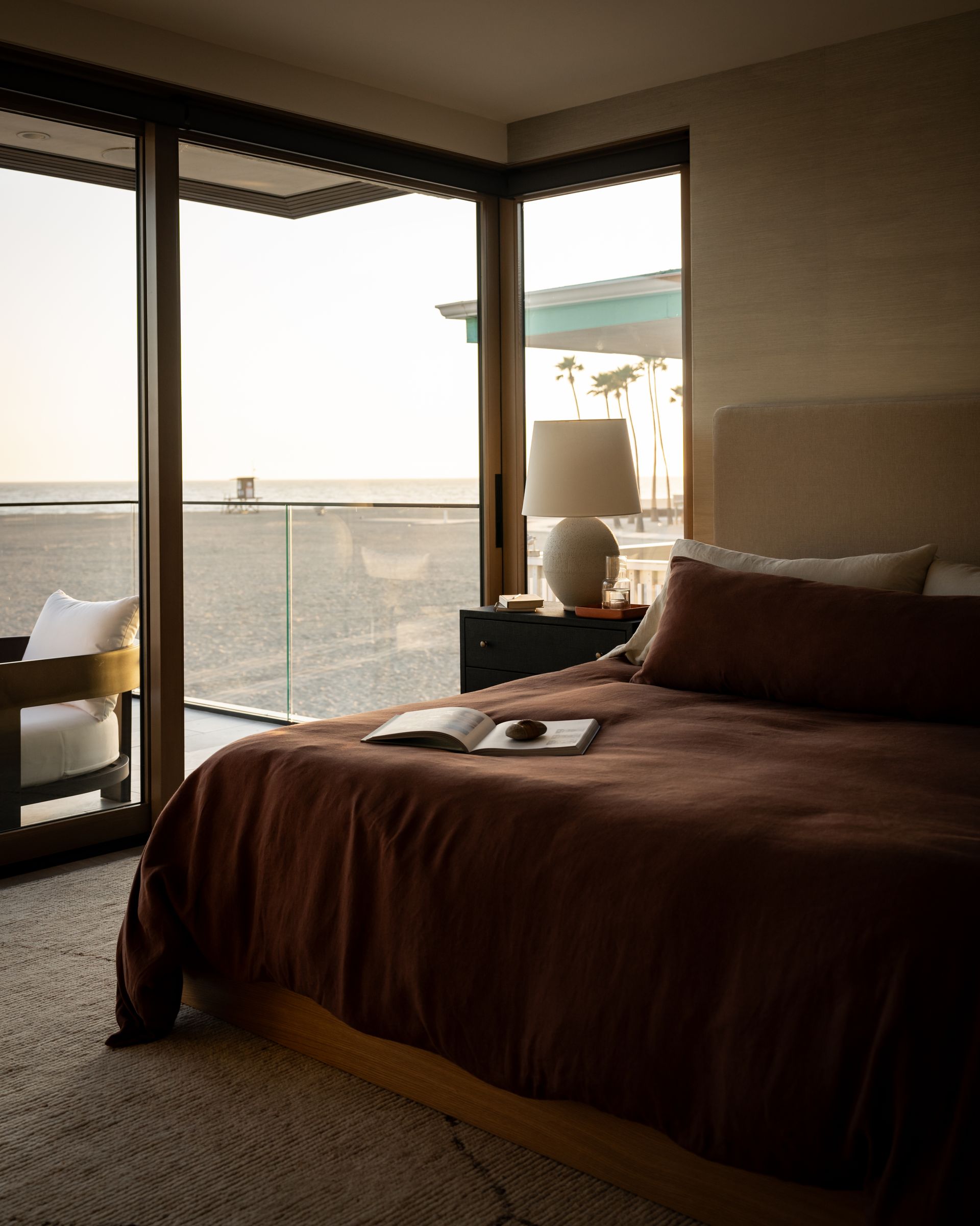 A bedroom with a dark brown comforter, a bedside lamp, and floor-to-ceiling windows overlooking a beach at sunset.