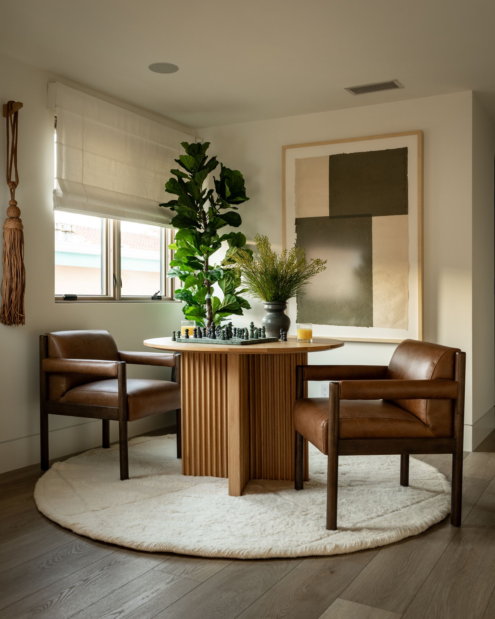 A round wooden table with a chess set and plant, flanked by two brown leather armchairs on a round rug in a bright room.