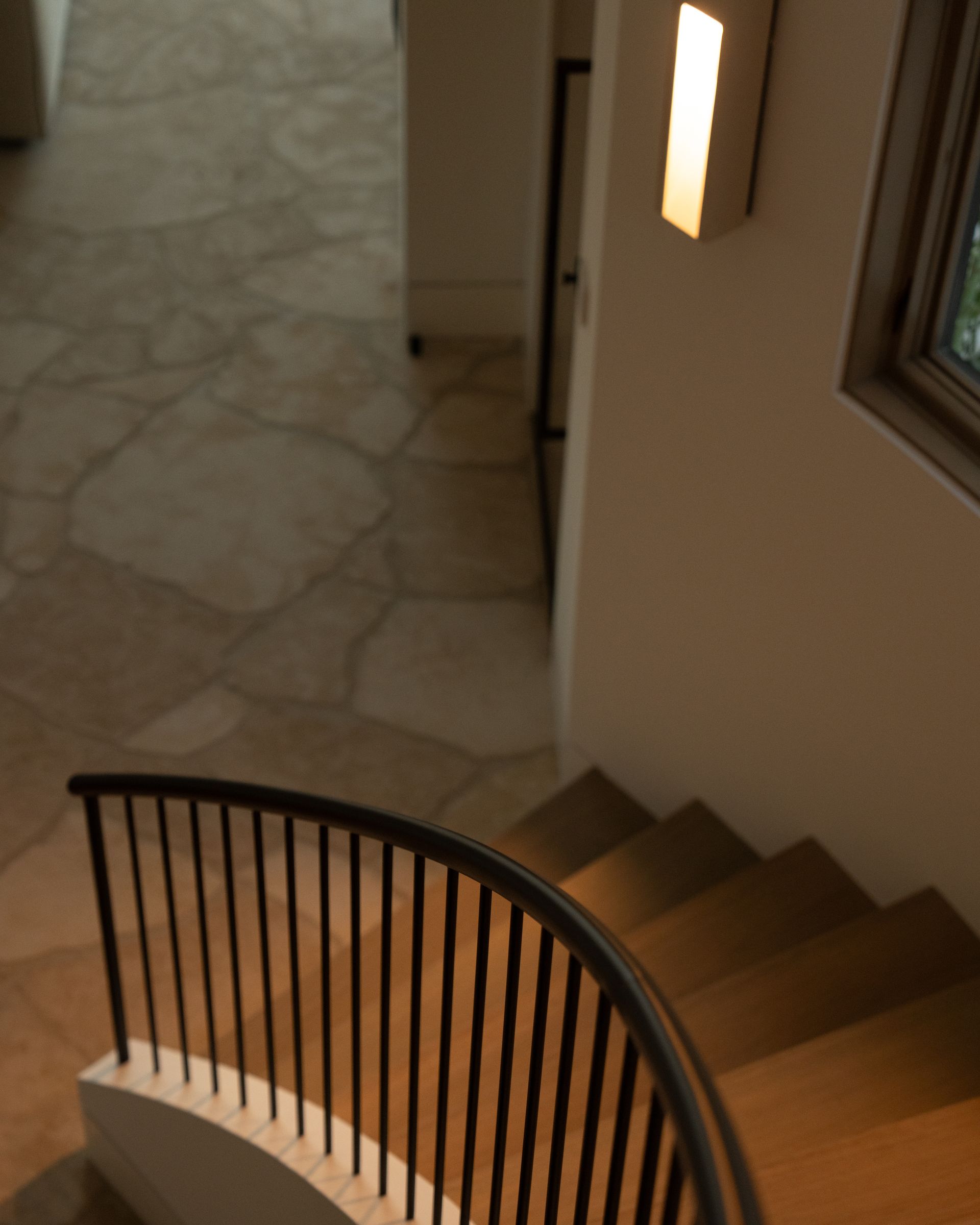 A downward angle of a curved staircase with wooden steps and a black metal railing, leading to a stone-tiled hallway.