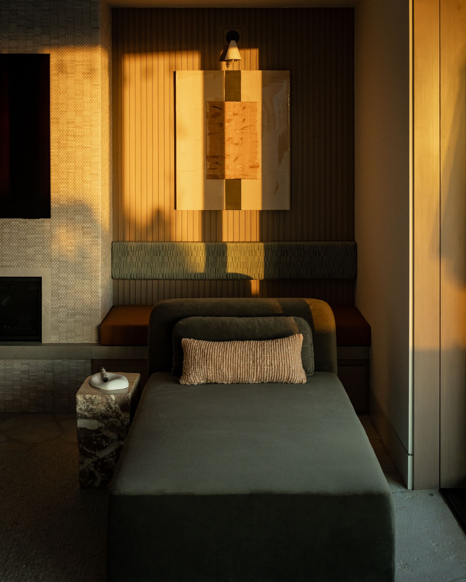 A dark gray daybed sits in a modern room, illuminated by warm sunset light, with a patterned pillow and wall art above.