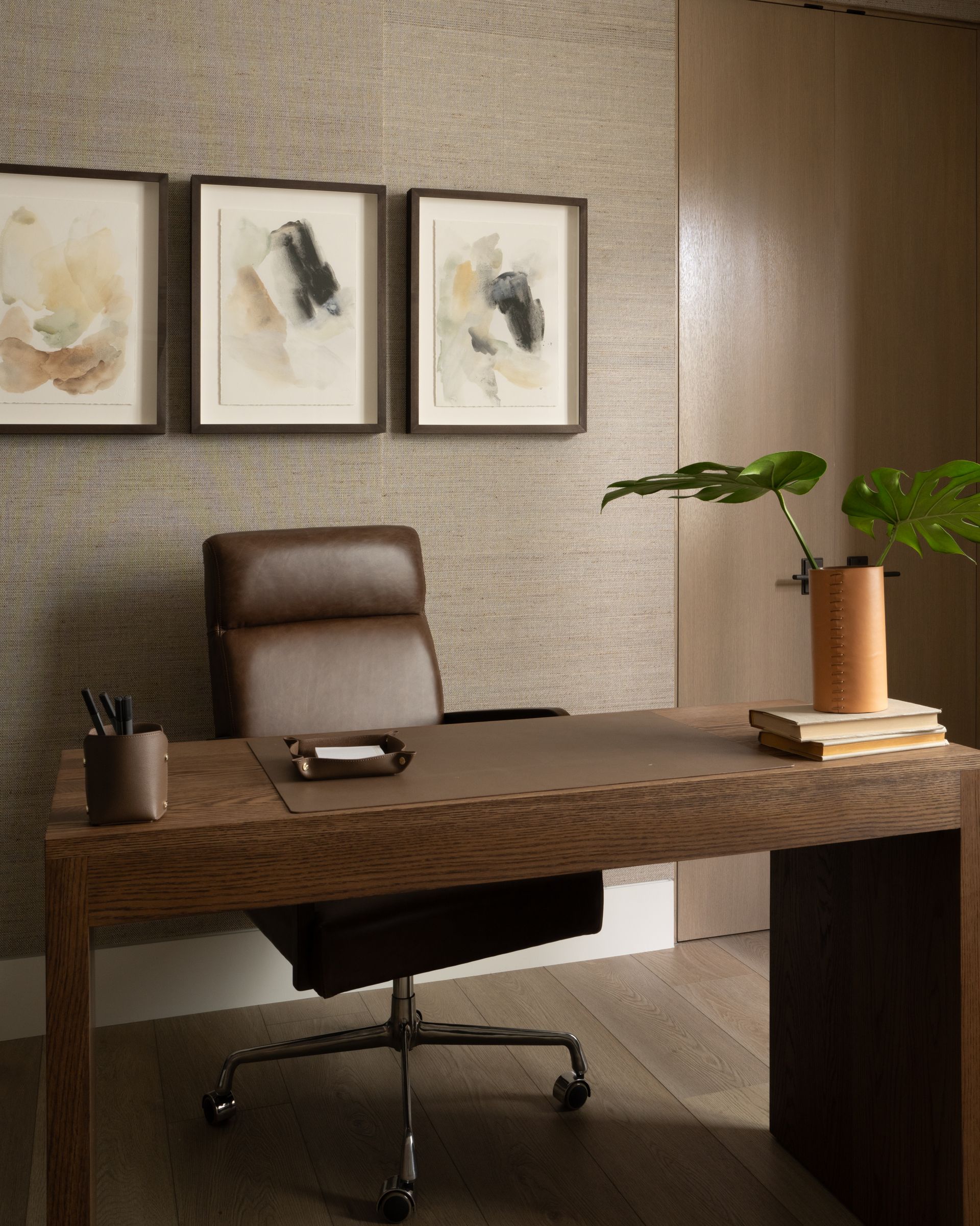 A modern desk and leather chair in a neutral-toned office with three framed abstract prints on a textured wall.