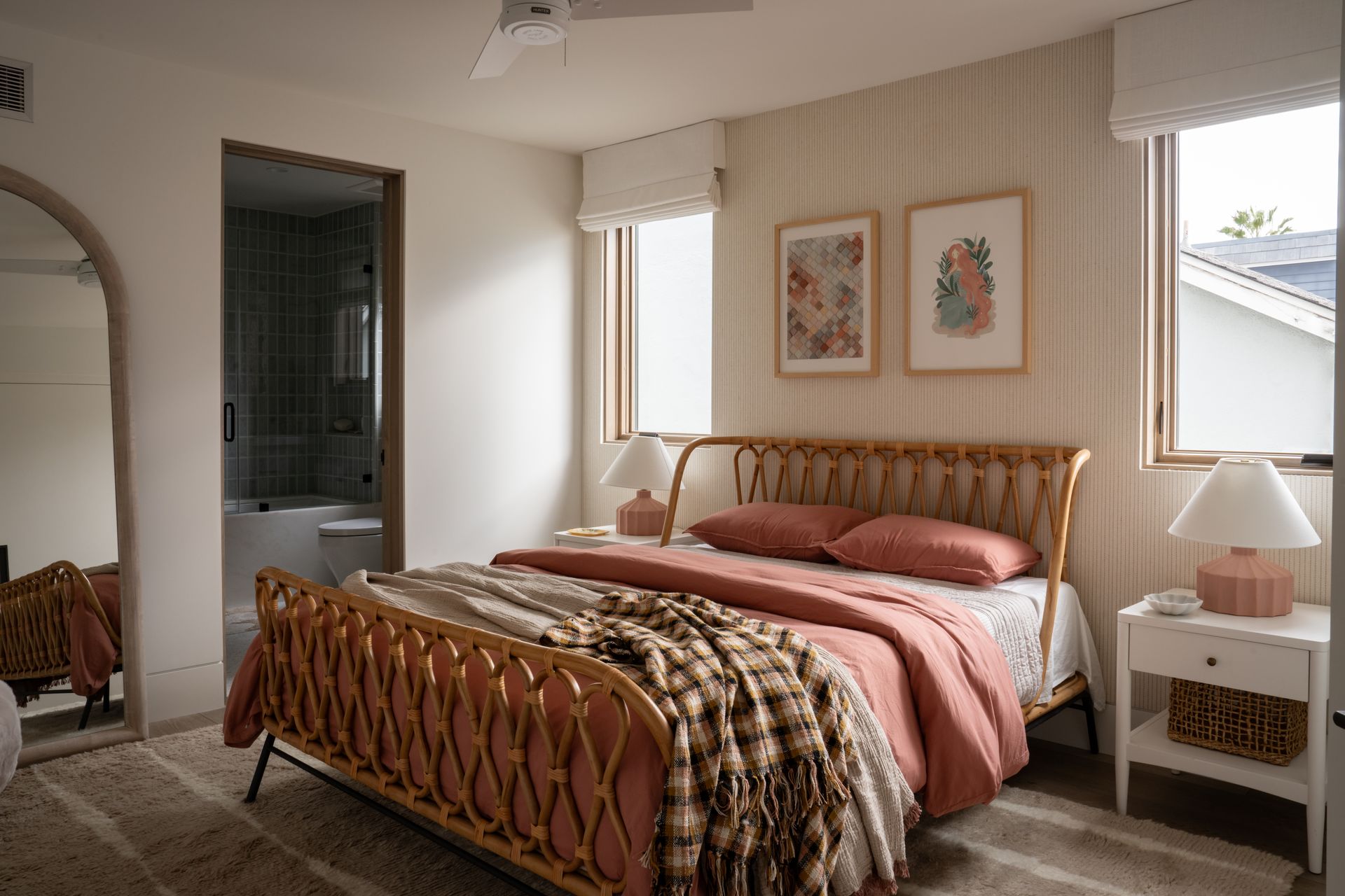 A cozy bedroom with a rattan bed, terracotta bedding, a plaid throw, two framed prints, and matching bedside lamps.