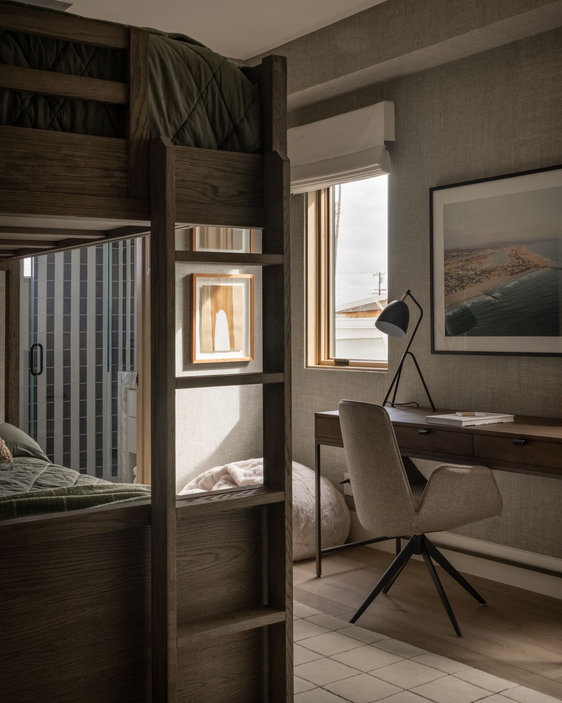 A bedroom featuring a wooden bunk bed, a desk with a lamp, a chair, and framed wall art in a sunlit, neutral-toned room.