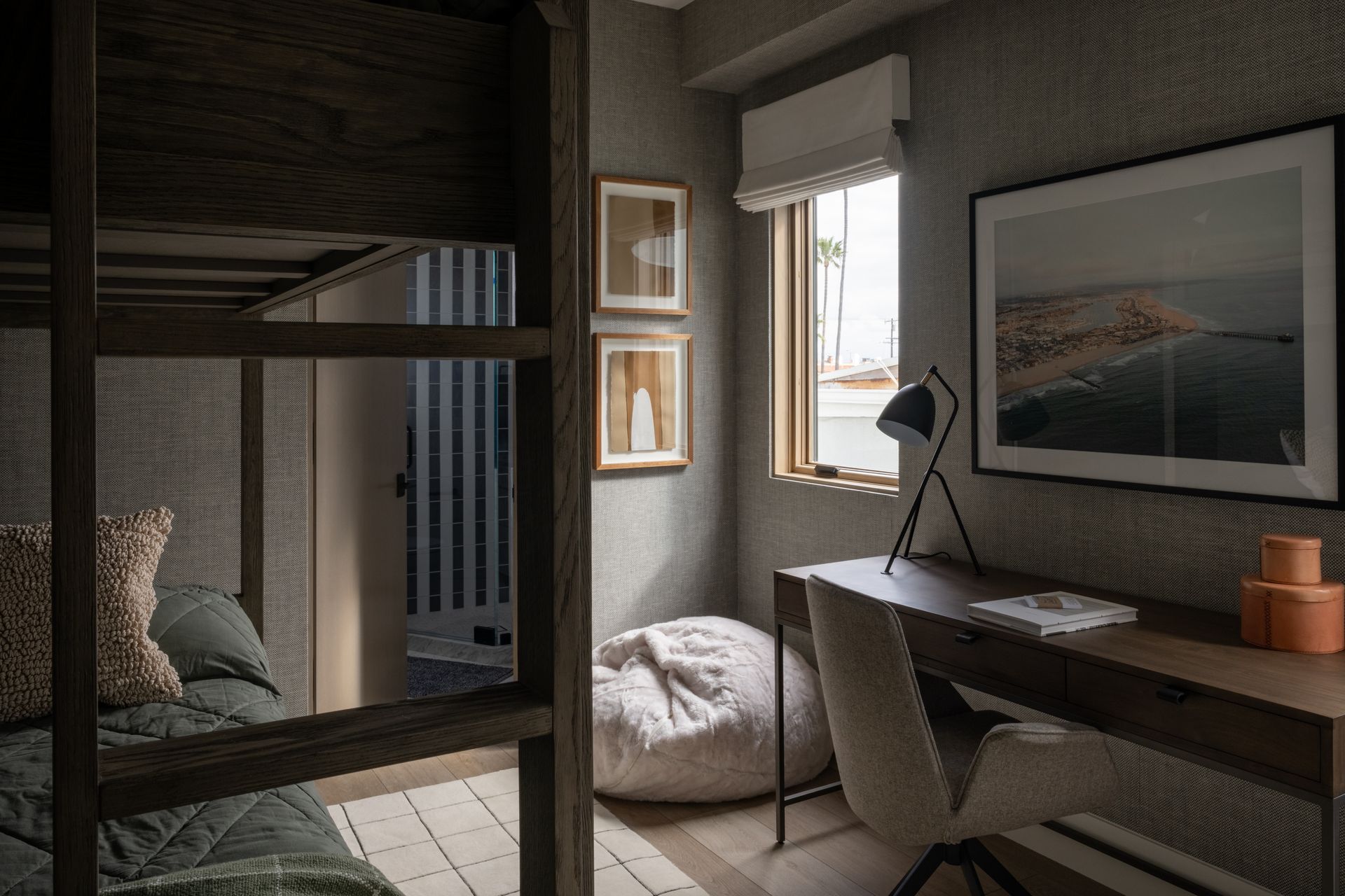 A cozy bedroom featuring a wooden bunk bed, a desk with a chair, a beanbag chair, wall art, and a small window with a shade.