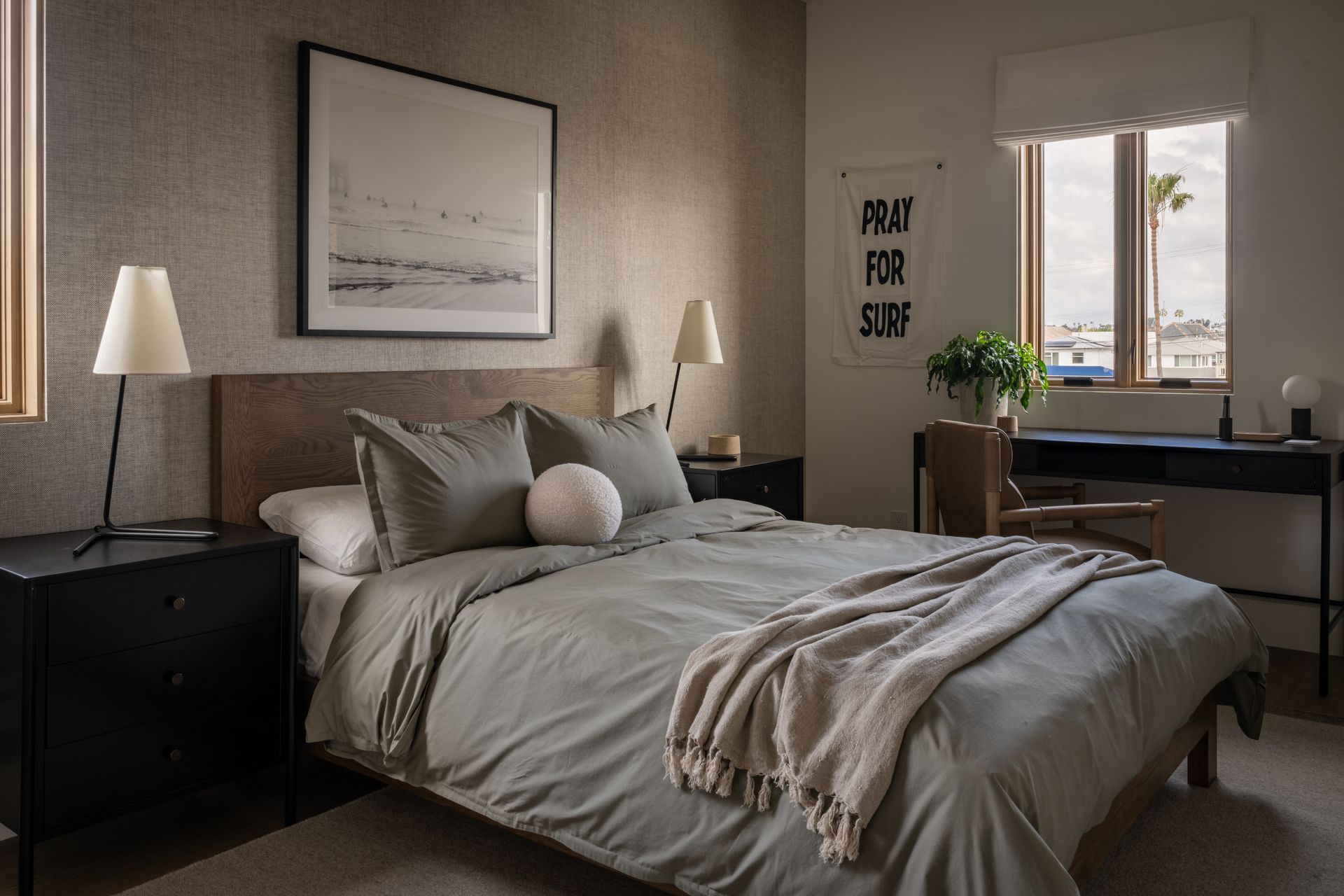 A modern bedroom featuring a sage green bed with a knit throw, black nightstands, a desk, and a framed beach photograph.