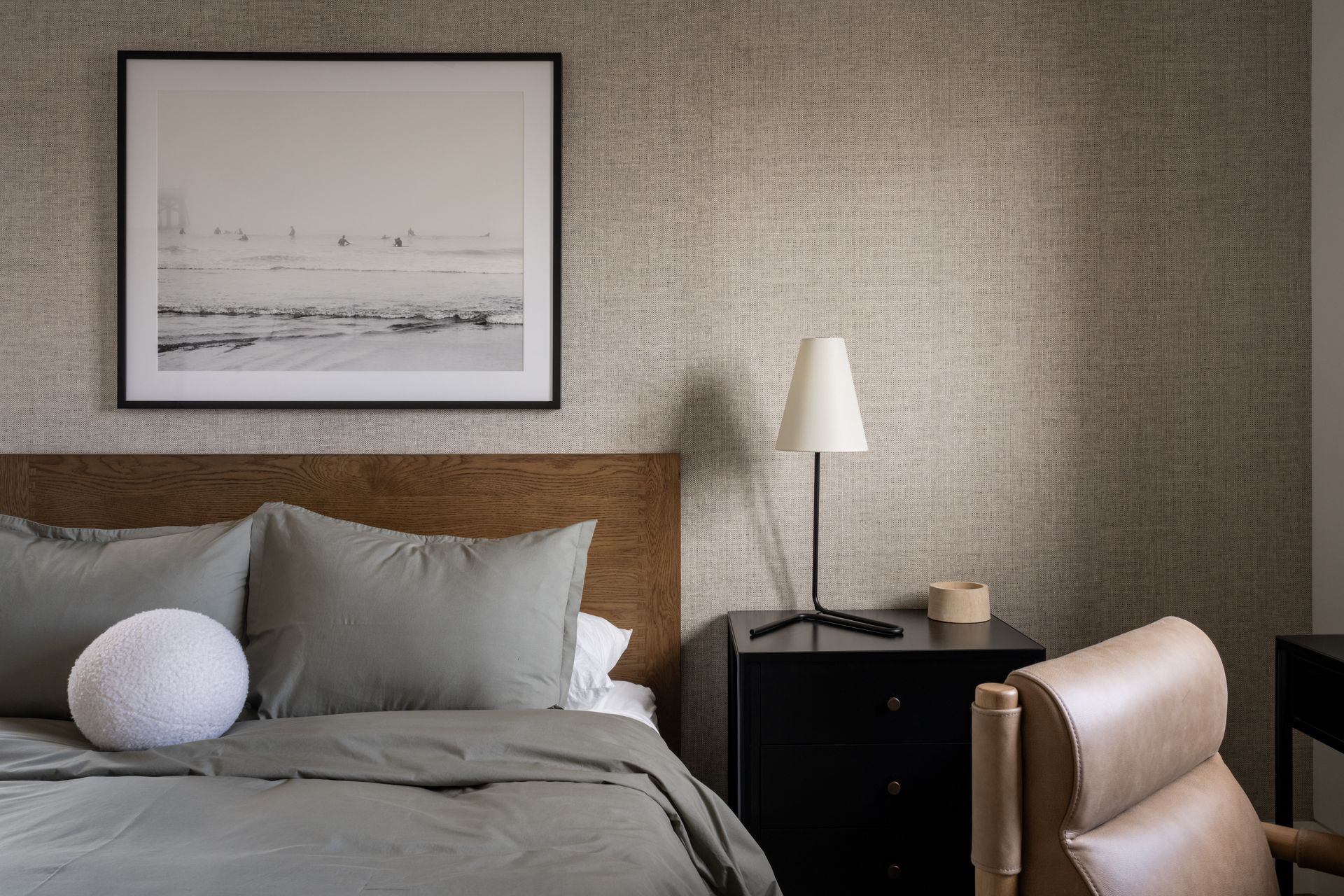 A bedroom with a wooden headboard, sage green bedding, a textured wall, black nightstand, lamp, and framed artwork.