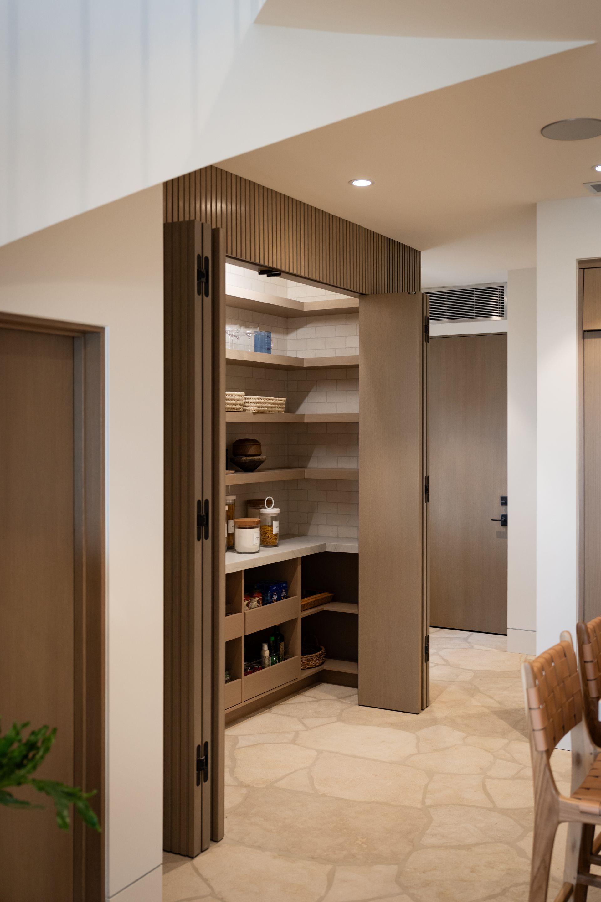 A corner walk-in pantry with light wood shelving, neutral stone flooring, and bifold doors in a modern home interior.