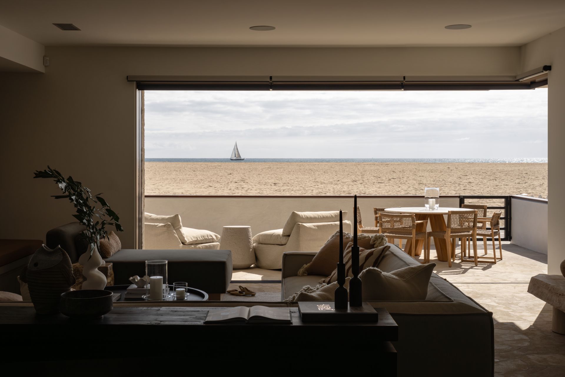 A modern living room opens onto a patio overlooking a sandy beach and the ocean on a bright, cloudy day.