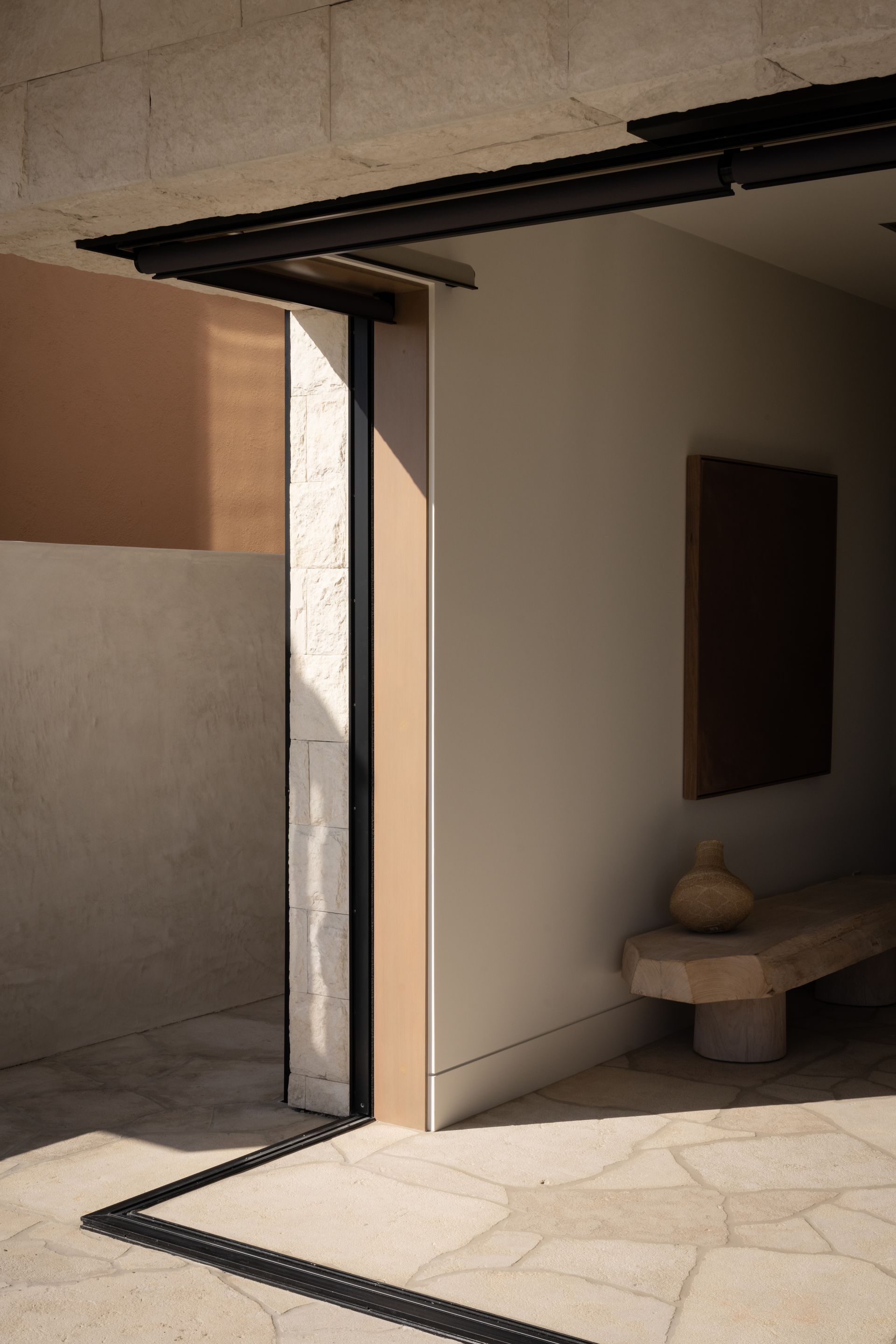 A sunlit outdoor entryway with beige stone floors, a minimalist bench, and a dark, modern sliding door frame.