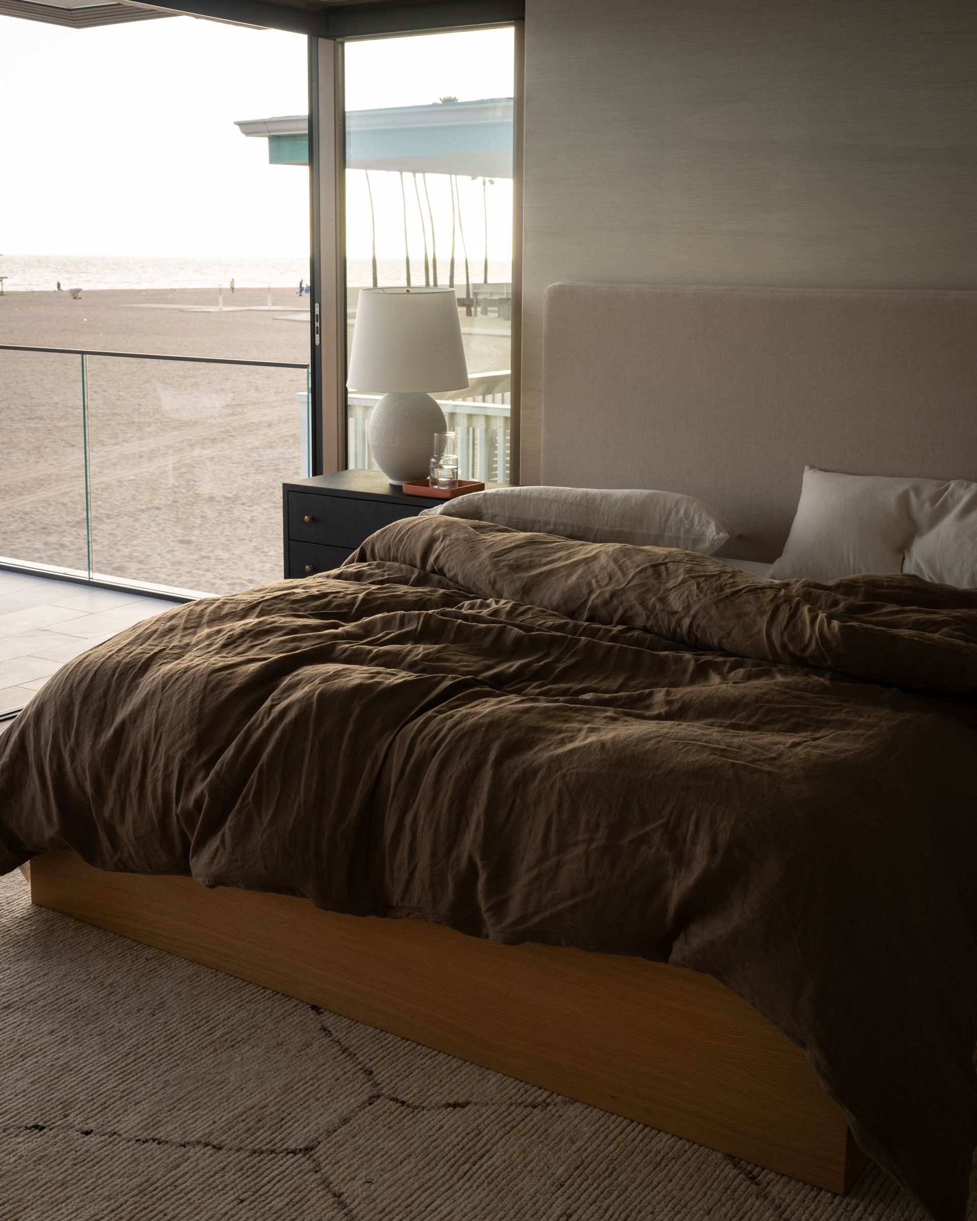A bed with a dark brown duvet sits in a room with floor-to-ceiling windows overlooking a sandy beach.