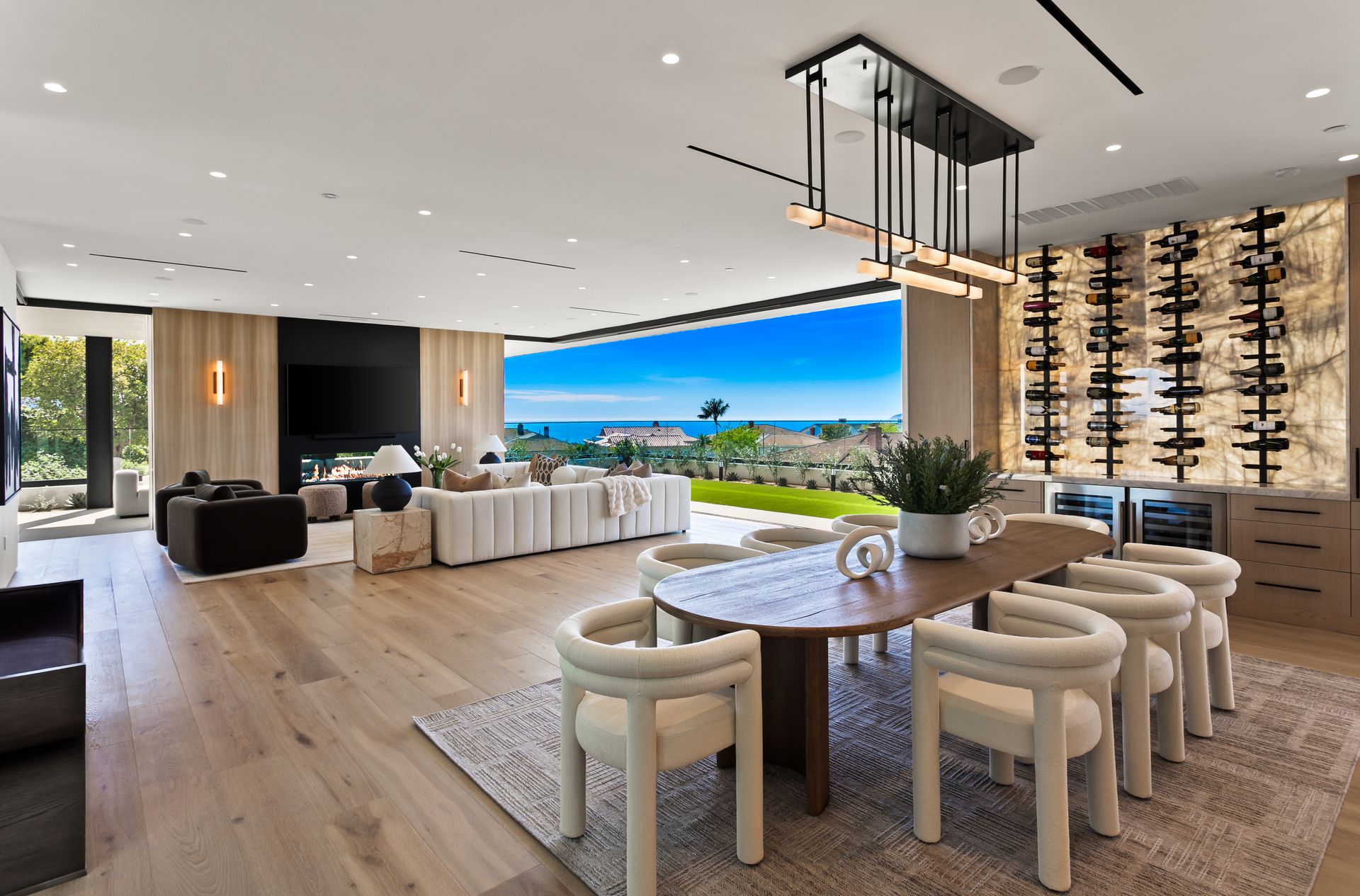 Modern open-plan living room with a backlit wine rack, sleek cabinets, dining table, and a scenic outdoor garden view.