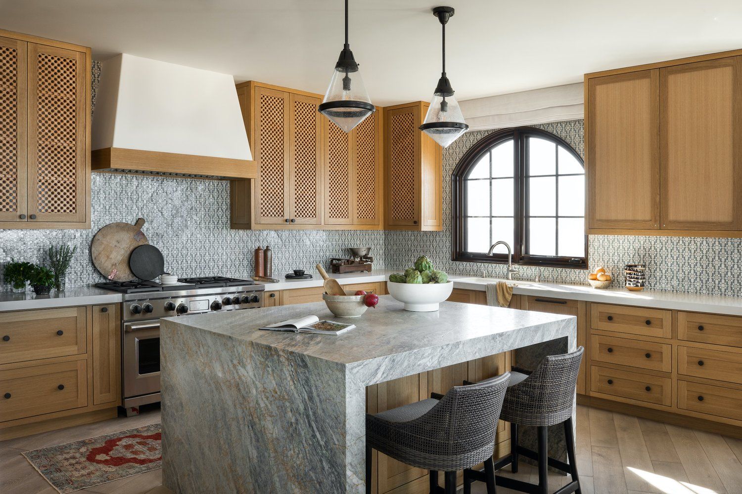 A bright kitchen with oak cabinetry, a stone-topped island, two gray stools, a white range hood, and an arched window.