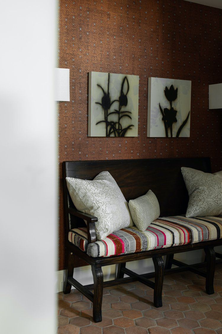 A dark wooden bench with striped fabric and patterned pillows sits against a textured brown wall with two floral prints.