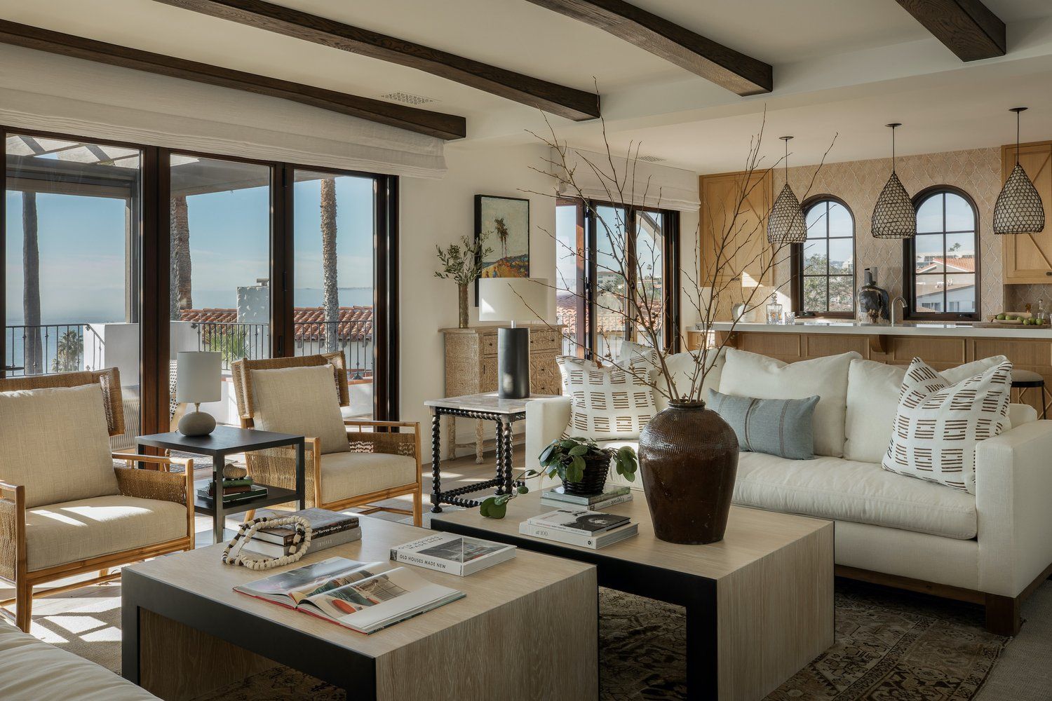 A bright, modern living room with a beige sofa, two armchairs, wooden coffee tables, and large windows overlooking the sea.