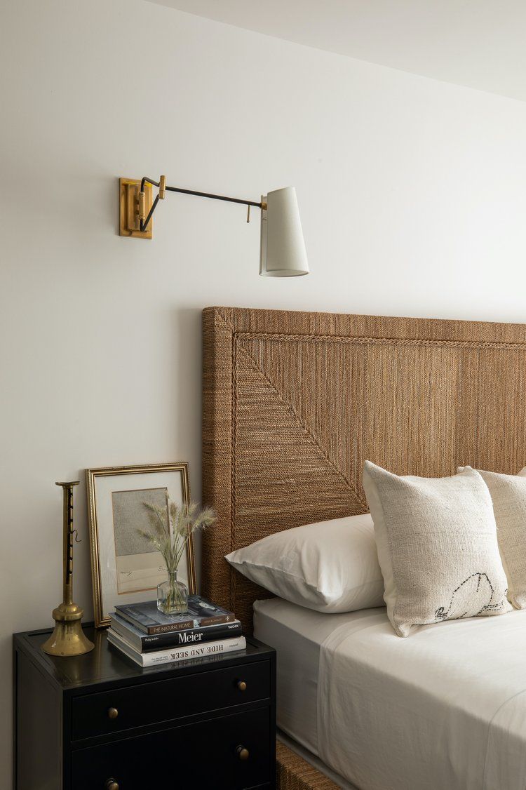A bedside setup featuring a woven headboard, a dark nightstand with books and a lamp, and a wall-mounted brass swing lamp.