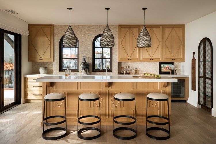 A kitchen with light wood cabinets, a white island with four stools, arched windows, and three woven pendant lights.