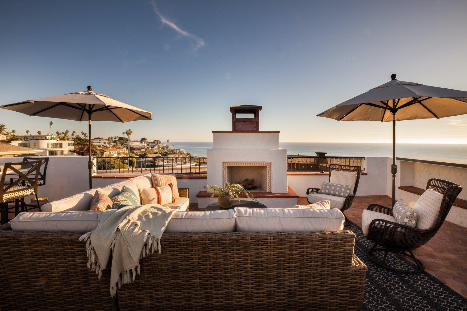 A scenic rooftop patio featuring outdoor furniture, two umbrellas, and a fireplace overlooking the ocean at sunset.