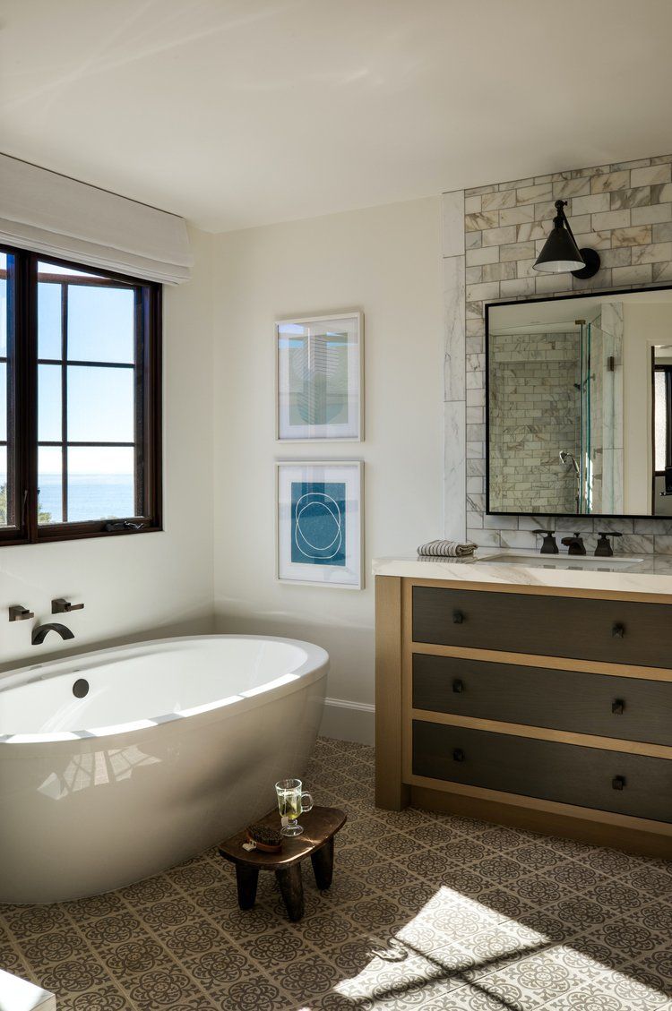 A modern bathroom with a freestanding white tub, a dark wood vanity, stone accent wall, and a window overlooking the ocean.