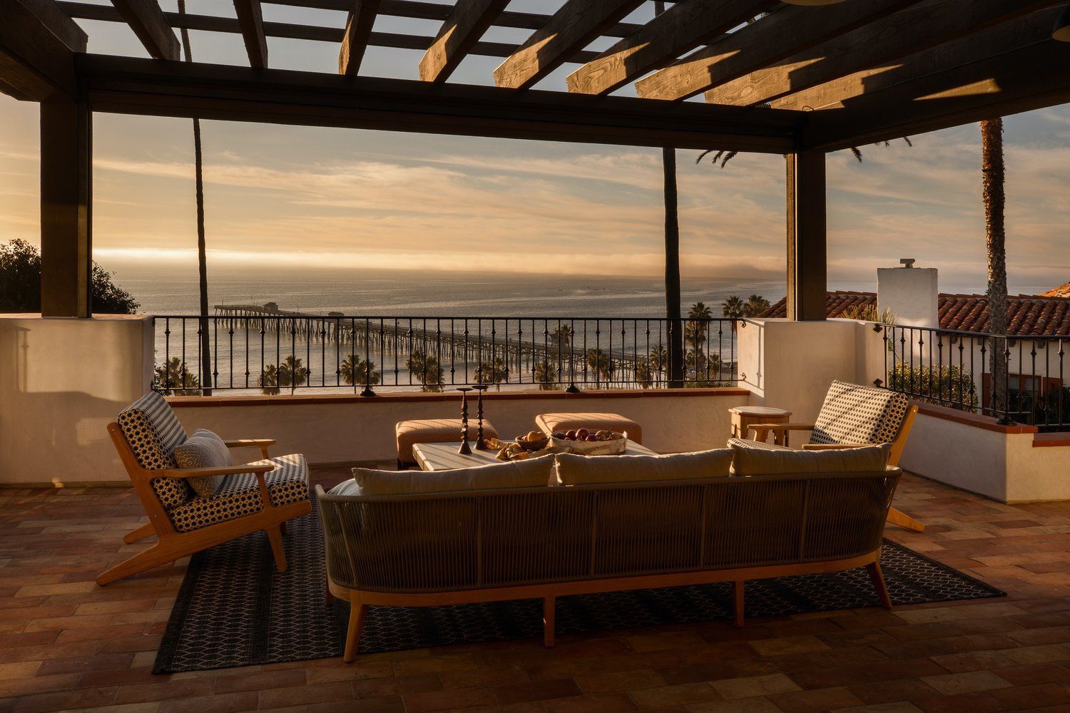A patio lounge area with a sofa, chairs, and a wooden pergola overlooking a coastal pier at sunset.