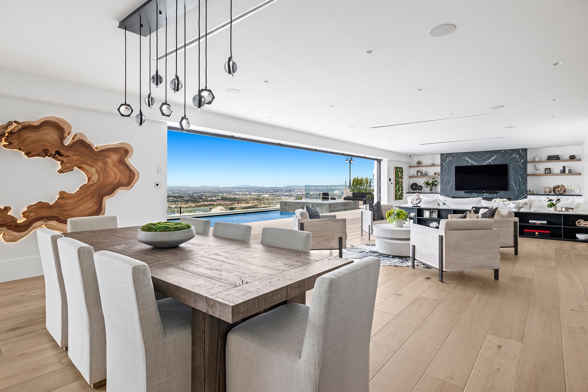 Modern open-plan living room with a long dining table, light wood floors, and floor-to-ceiling views of a pool and city.