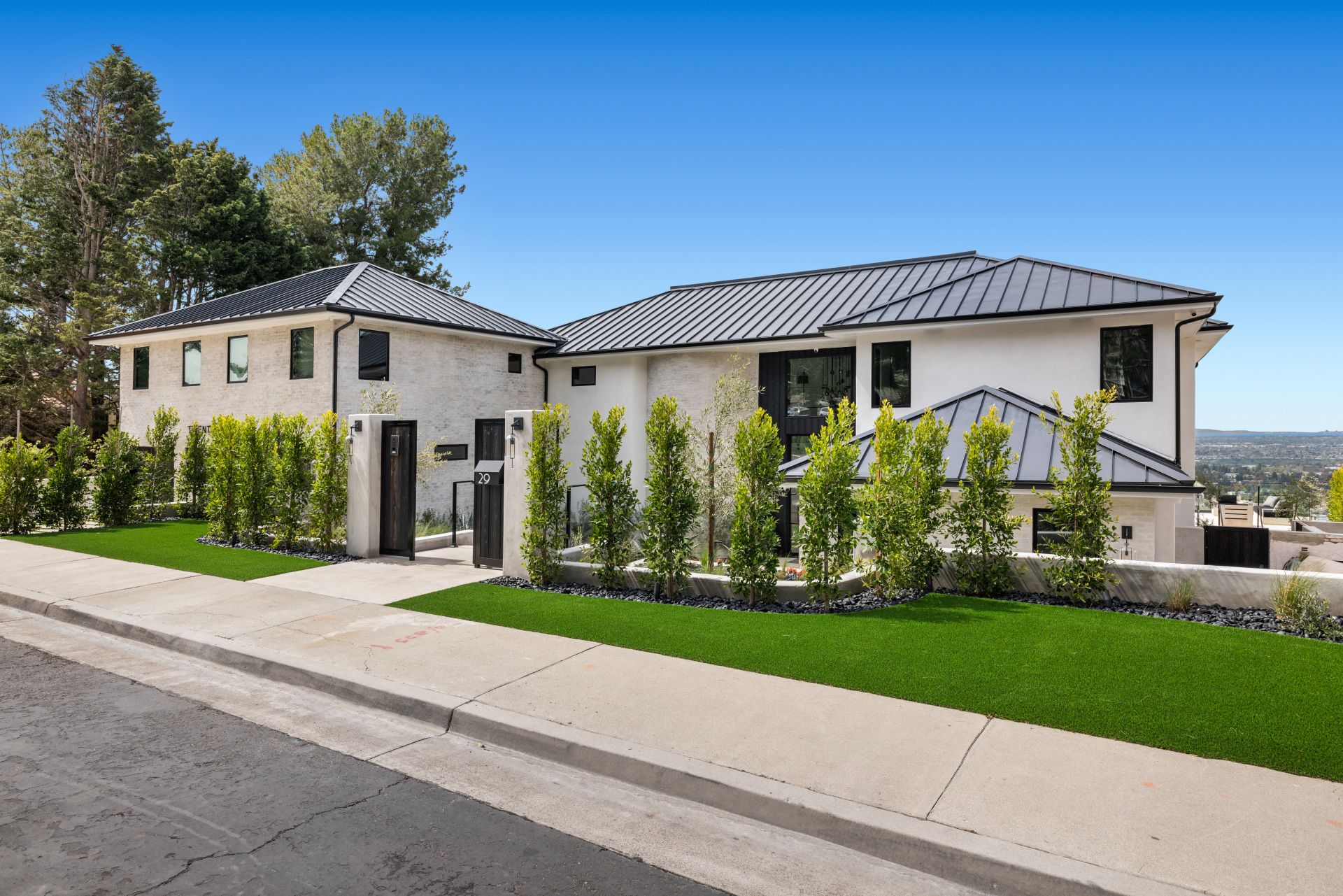 A modern white house with a dark metal roof, surrounded by a manicured hedge and green lawn under a clear blue sky.