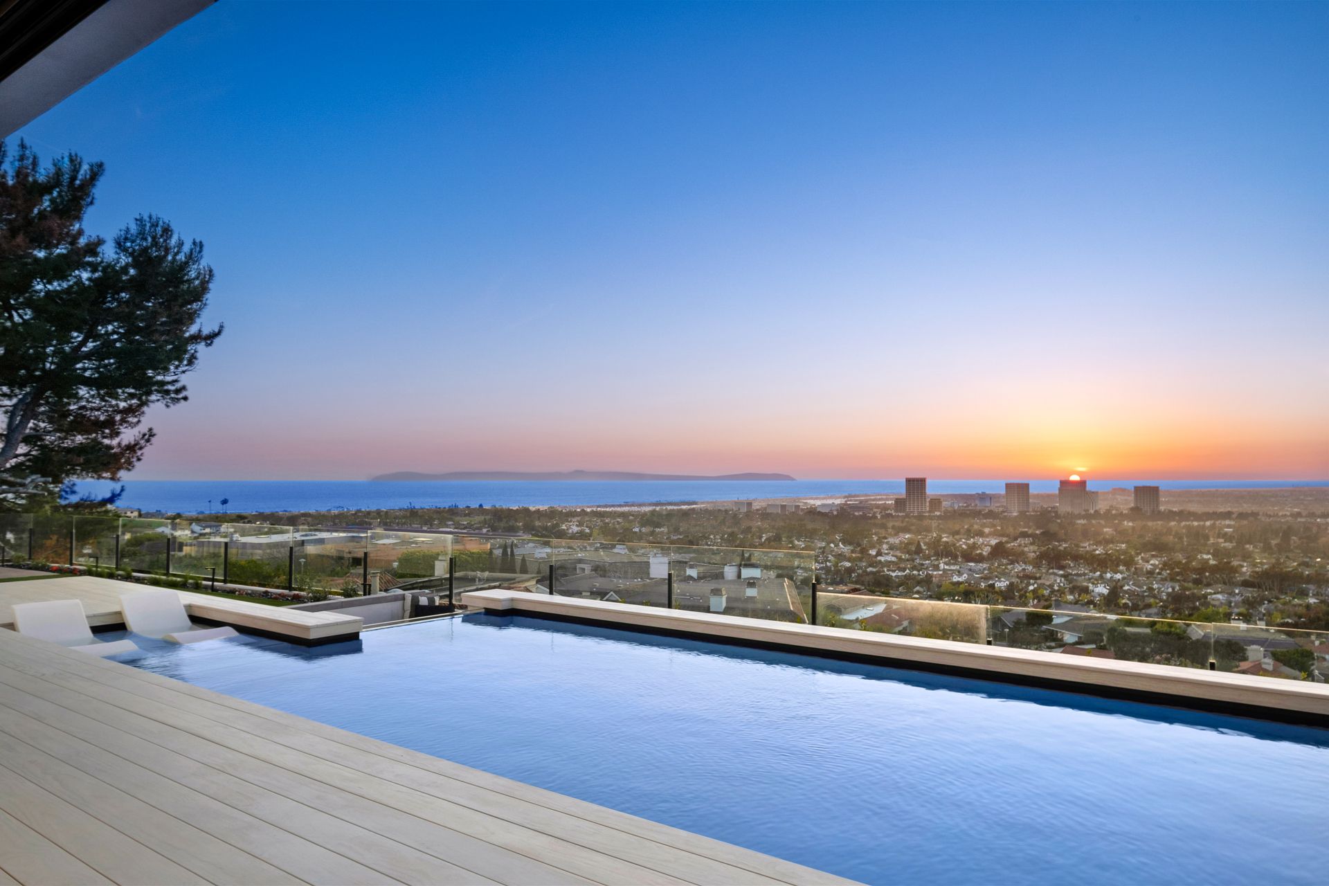 A modern infinity pool on a wooden deck overlooks a city skyline and ocean at sunset.