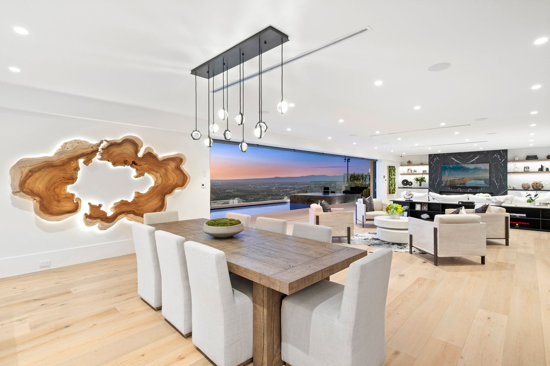 A dining room with a wooden table, white chairs, a statement wall piece with backlighting, and a scenic outdoor view.