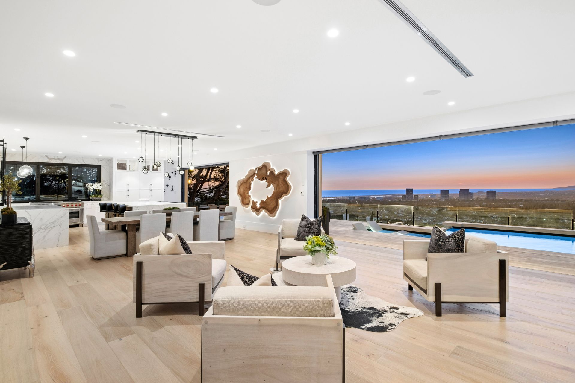 A luxury modern living room with neutral-toned seating, wood floors, and floor-to-ceiling glass walls overlooking a pool.