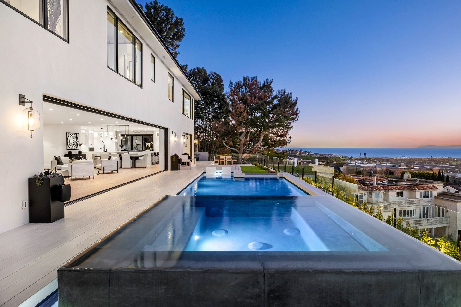 A modern white house with a large open patio and infinity pool overlooking a sunset view of a coastal city.