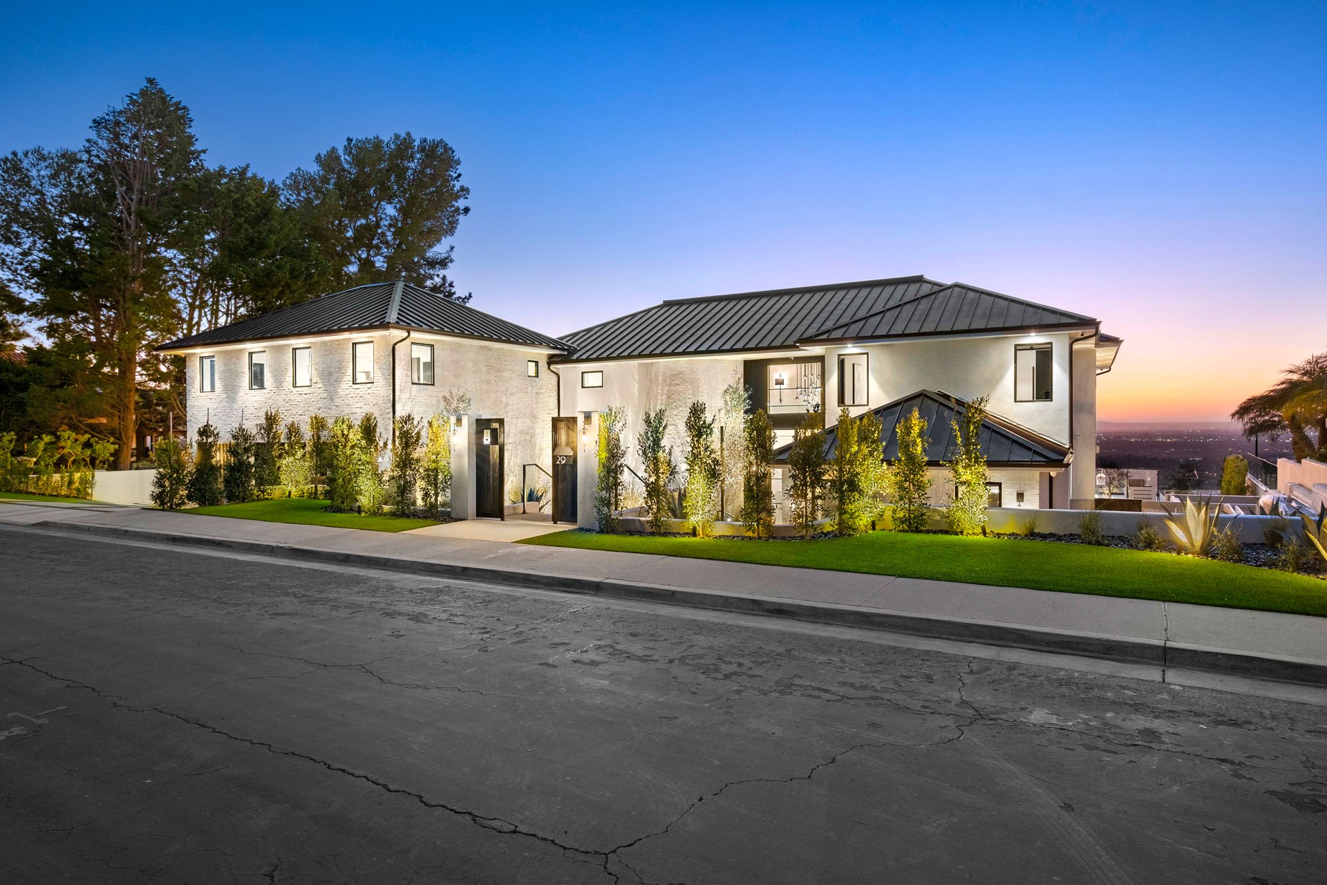 A modern, two-story white house with a dark, gabled roof illuminated at twilight, situated along a paved suburban street.