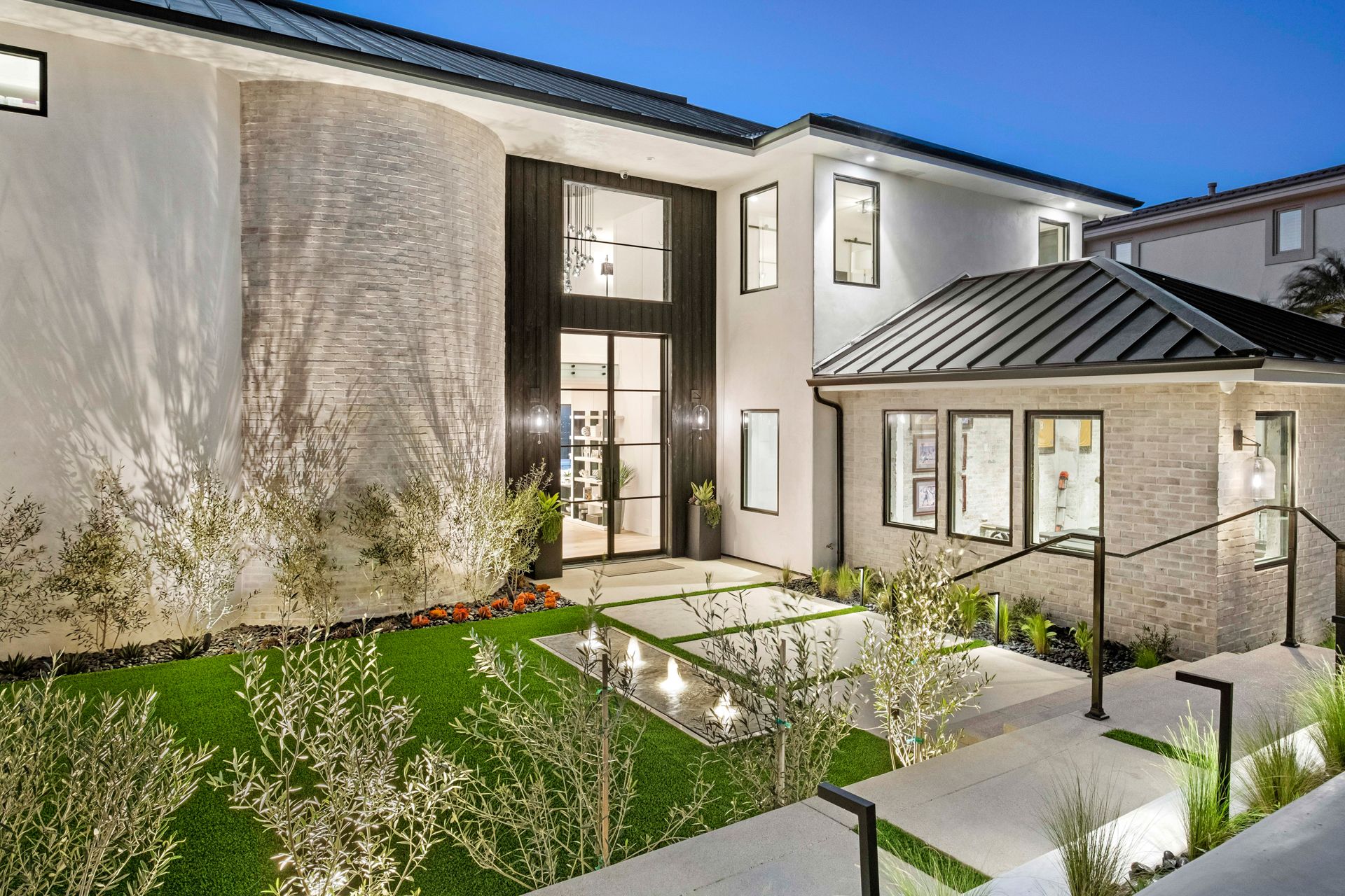 Modern two-story house with white walls, stone accents, black metal roof, and a lighted front walkway at twilight.