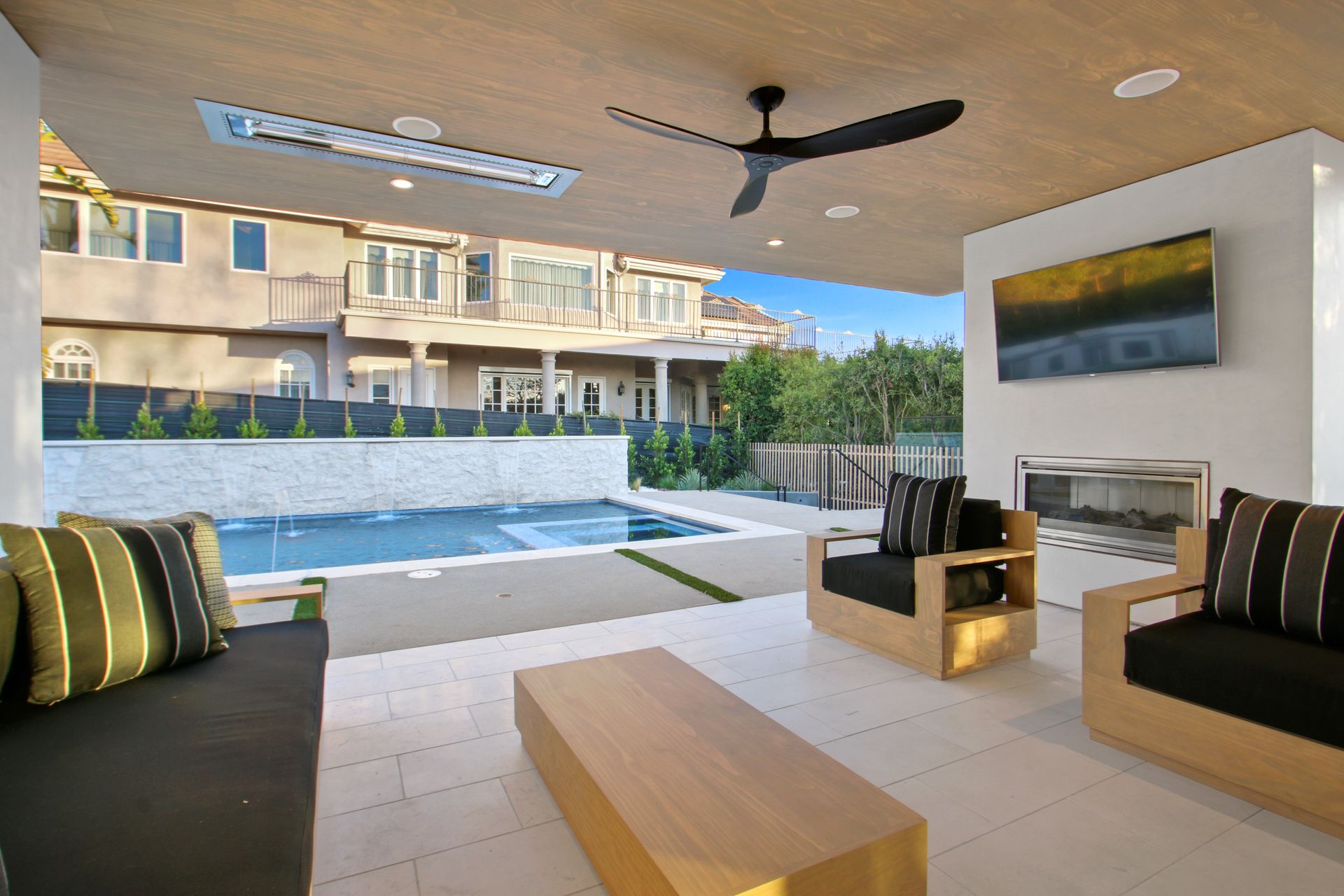 A modern covered patio with outdoor seating, a fireplace, and a television overlooking a private swimming pool.