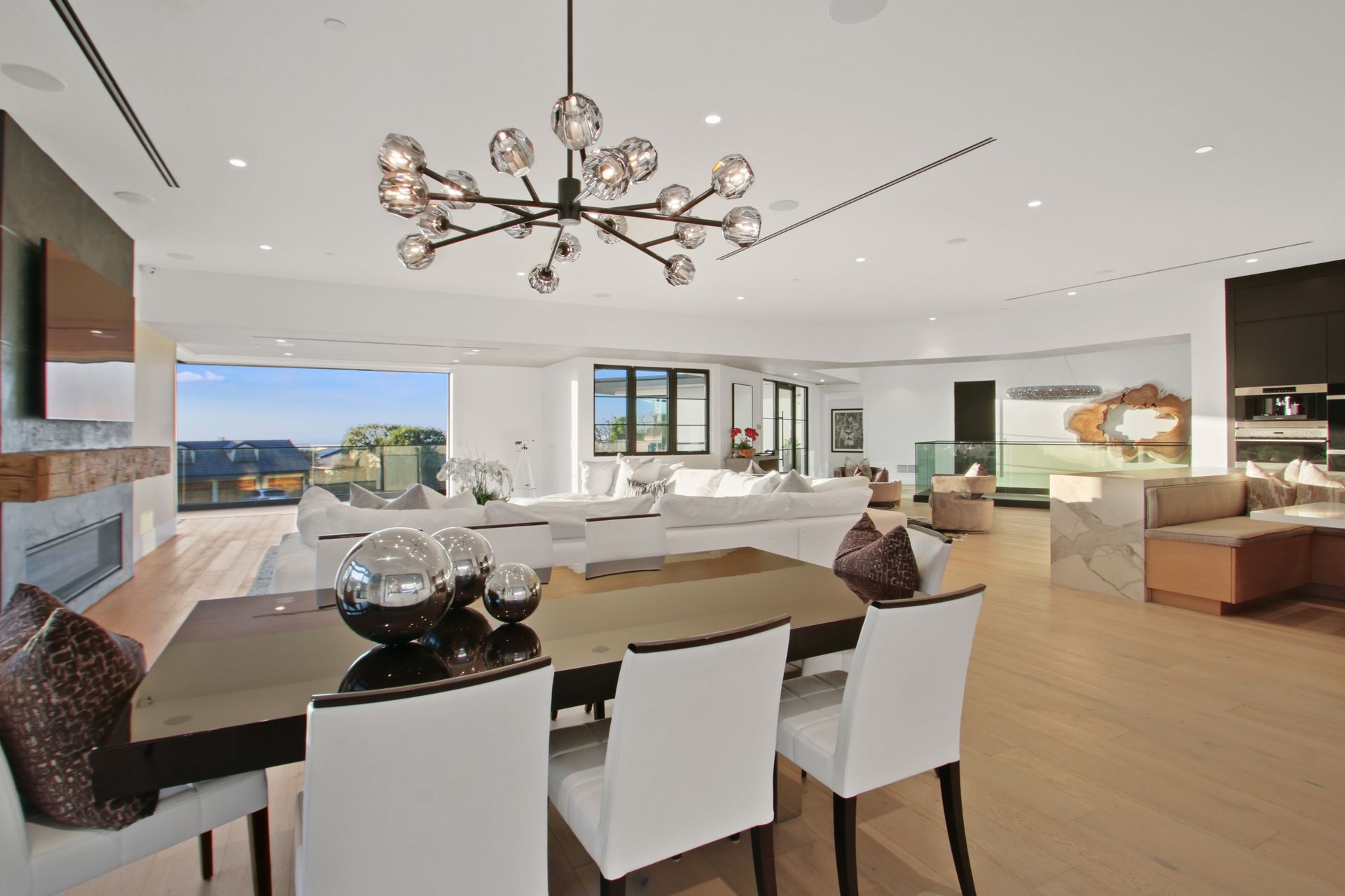 A modern, open-concept living and dining area with white chairs, a glass table, and a large chandelier.