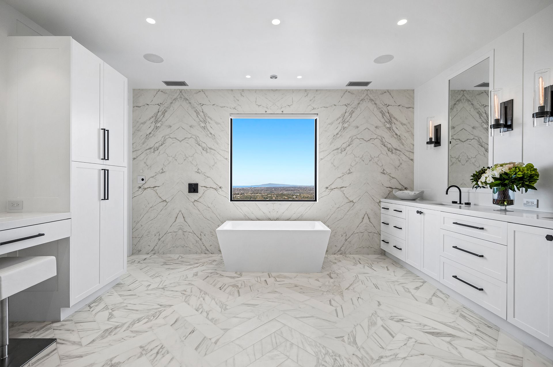 A modern, white-themed bathroom featuring marble walls and flooring, a central soaking tub, and a large window.