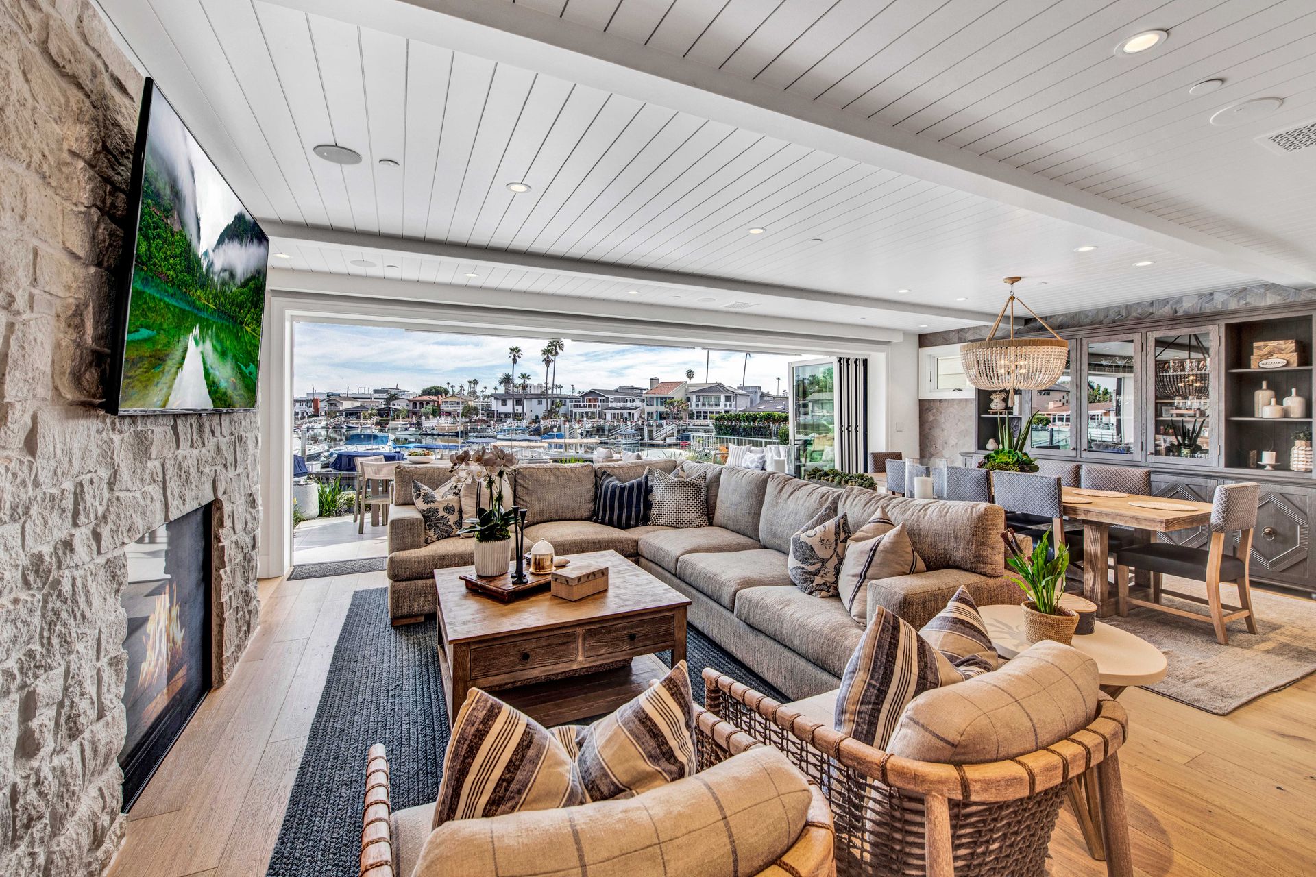 A living room with stone fireplace, neutral sectional sofa, and floor-to-ceiling glass doors opening to a harbor view.