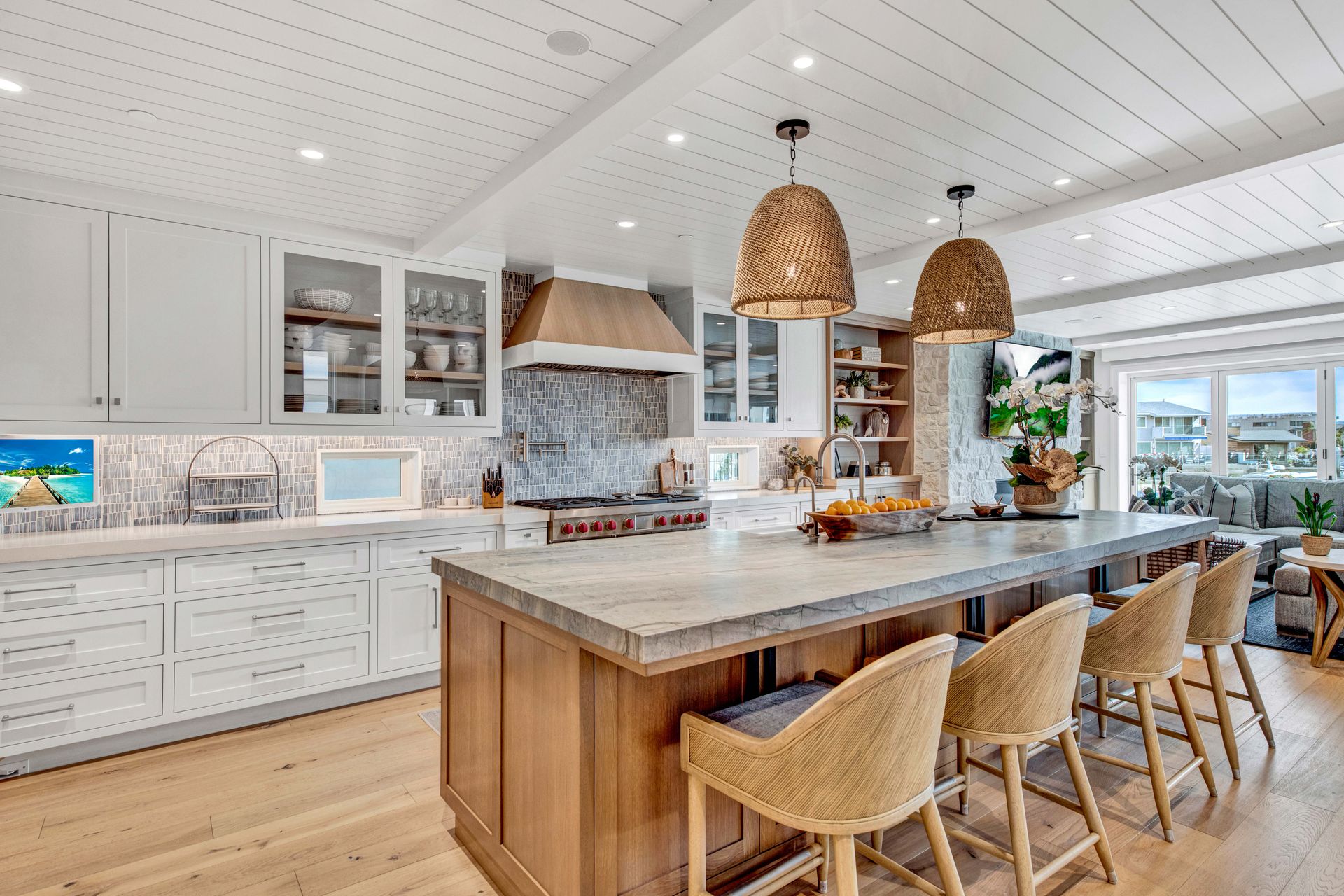 A bright, modern kitchen with white cabinets, light wood flooring, a large island with wicker stools, and pendant lights.
