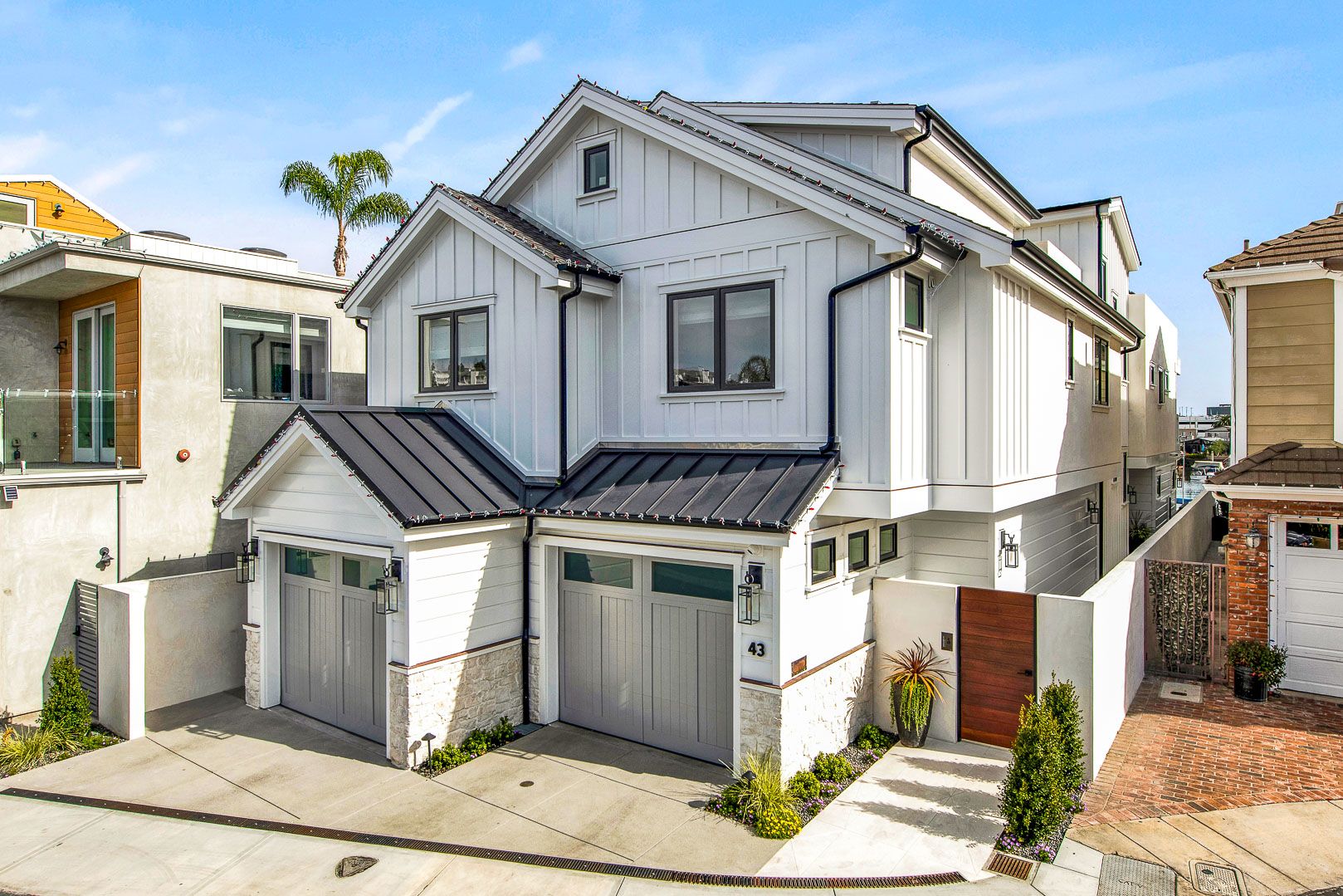 A two-story white farmhouse with two garage bays, stone accents, and a dark metal roof under a bright blue sky.