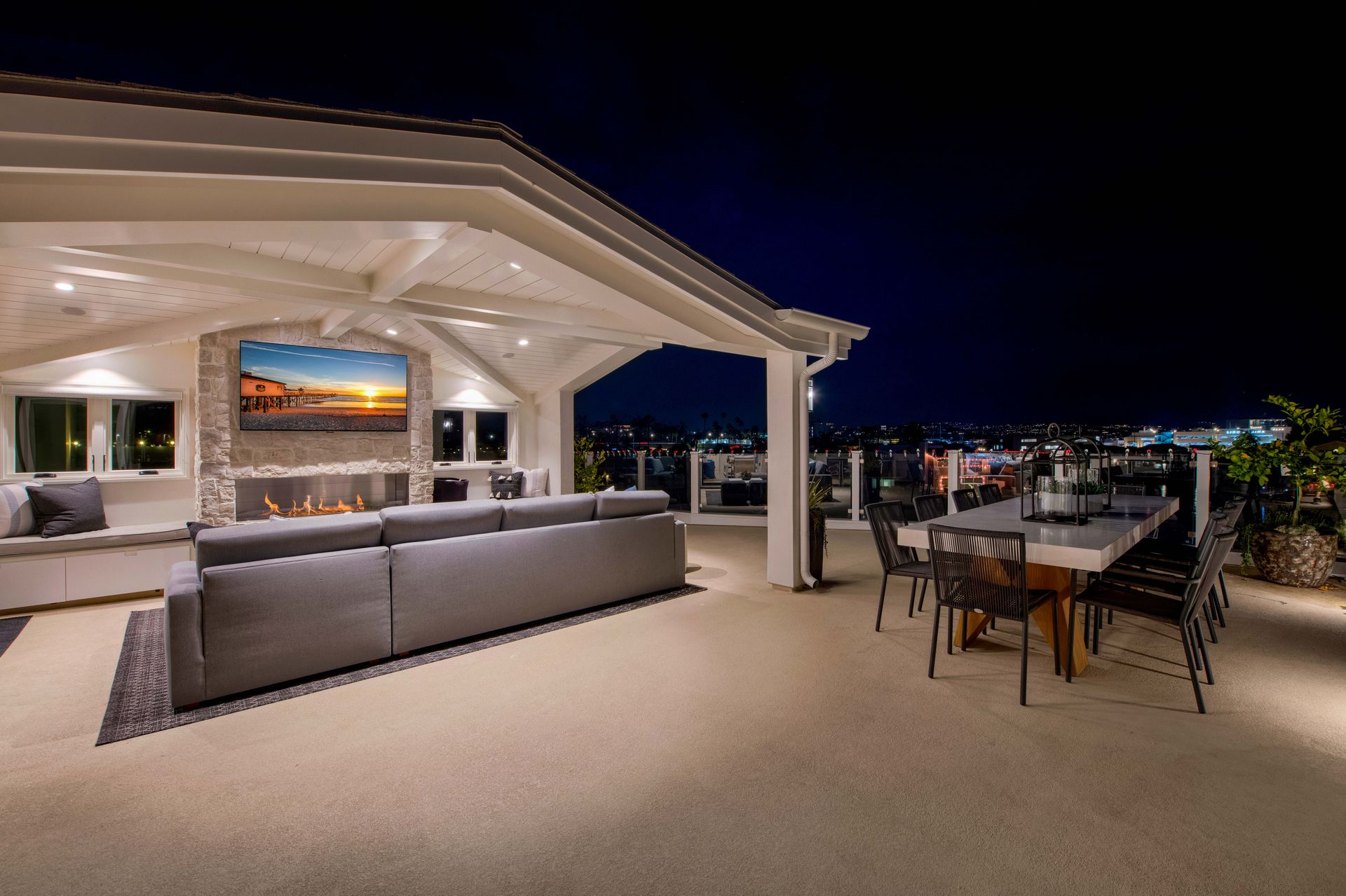 A modern outdoor patio at night featuring a covered lounge area with a fireplace, TV, gray sectional, and dining set.