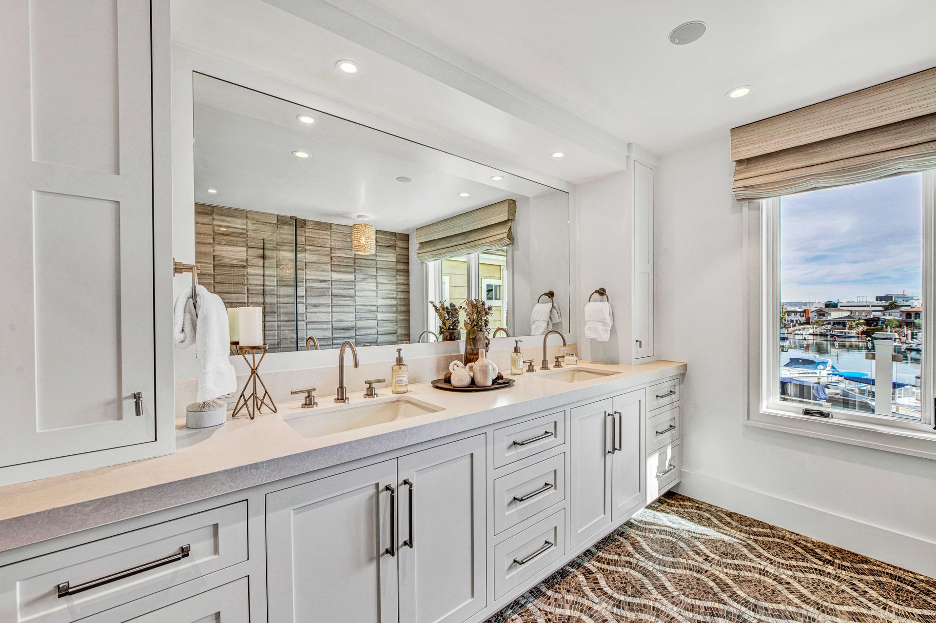 A modern, bright bathroom with white cabinetry, double sinks, a large mirror, and a window overlooking a marina.