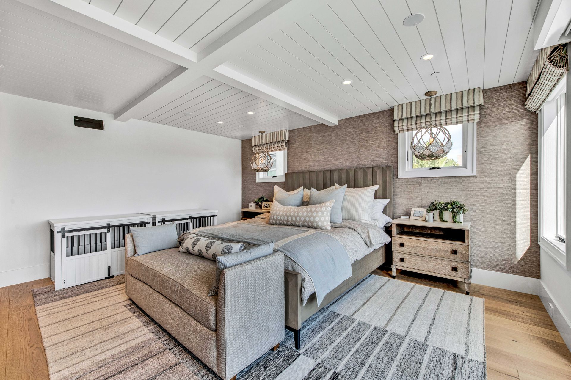 A bedroom with a grey tufted bed, white shiplap ceiling, wood nightstands, and patterned rug on light wood flooring.