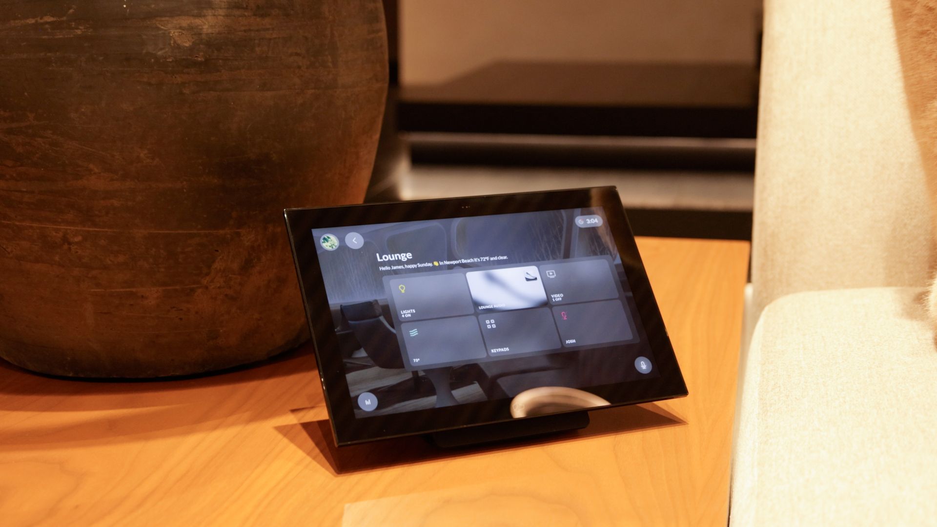 A smart home control tablet sits on a wooden surface next to a large, dark vase, showing a lighting management interface.